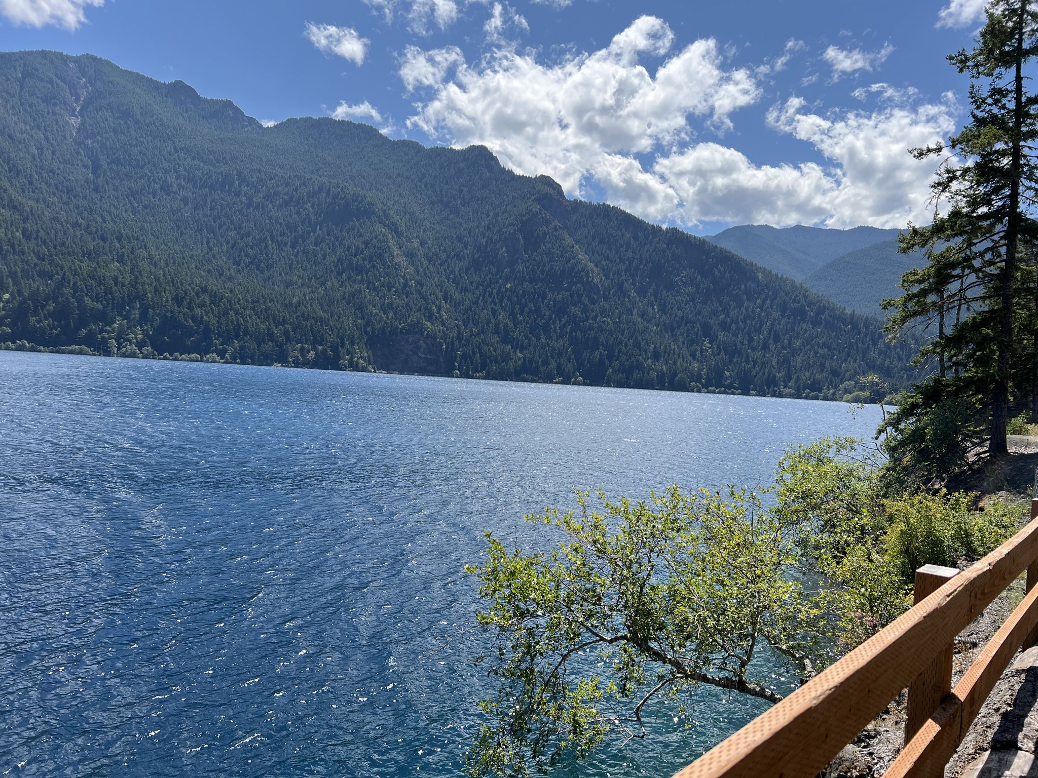 Vast blue lake with forested mountains under a bright, cloudy sky. - Spruce Railroad Trail