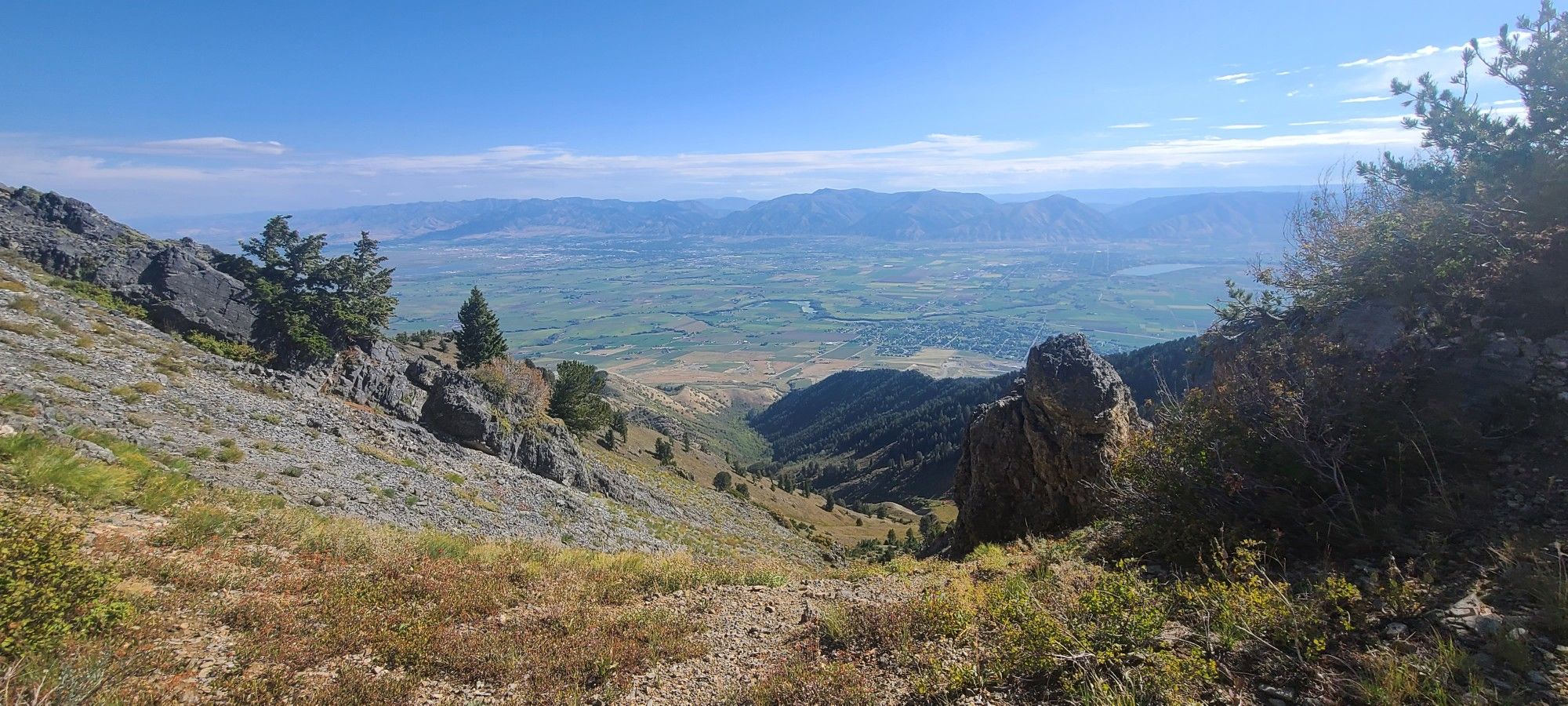 Sunny view down a rocky mountain slope to a wide green valley. - Wellsville Ridge Trail
