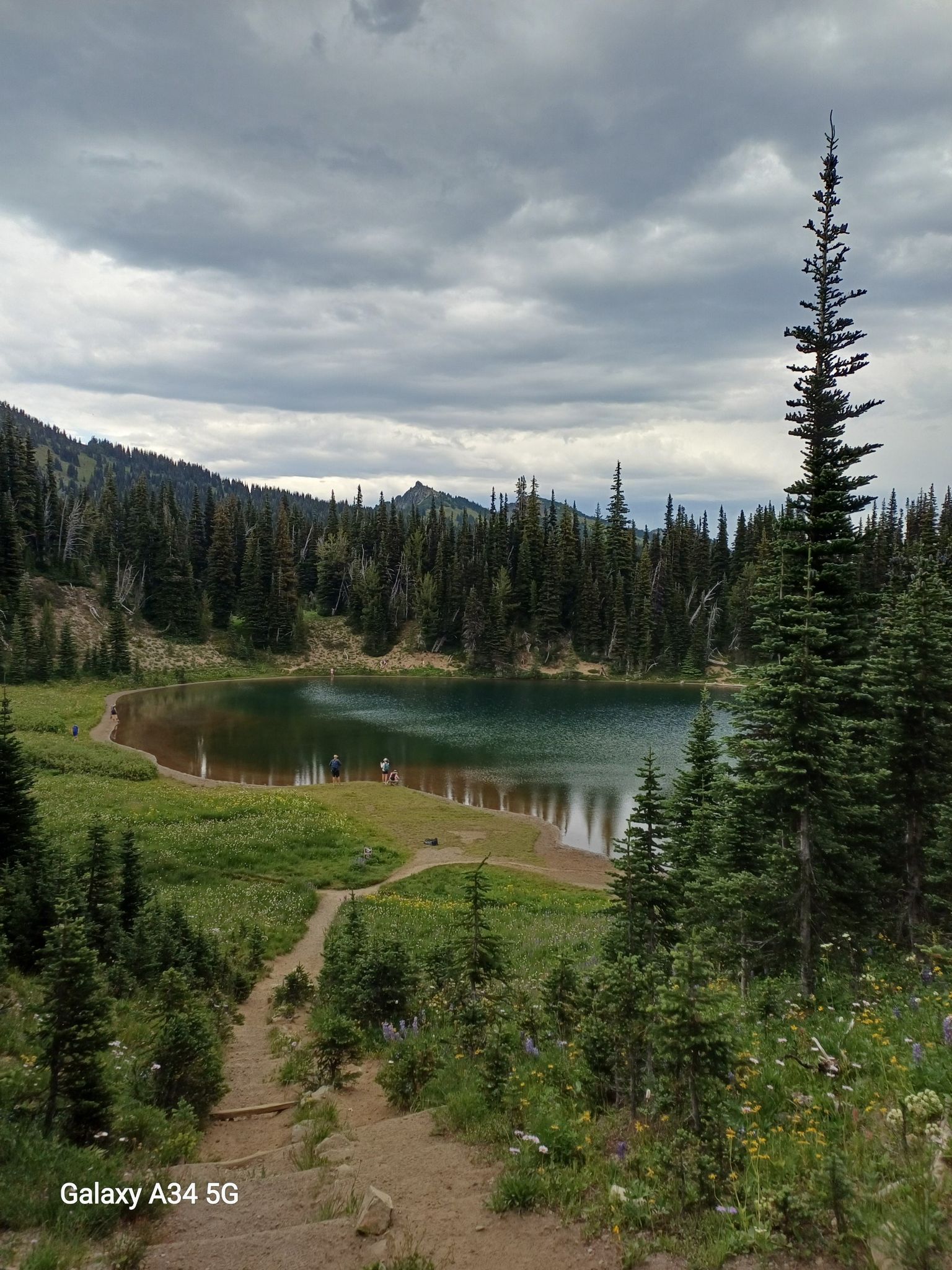 Dirt path descends to a lake with people, surrounded by evergreen trees under cloudy sky. - Shadow Lake Trail