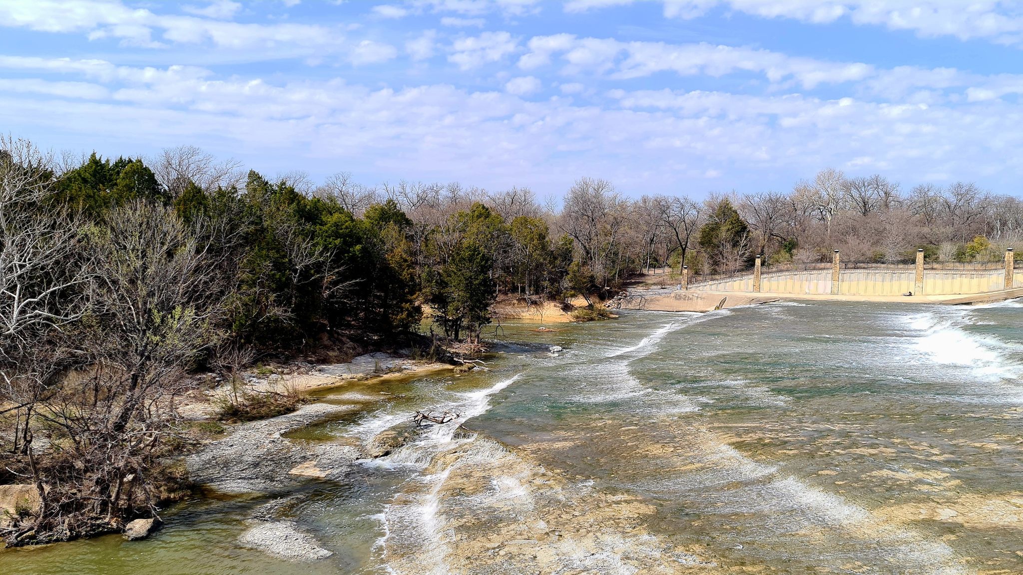 River rapids flow past bare and evergreen trees under a bright sky. - White Rock Lake Trail