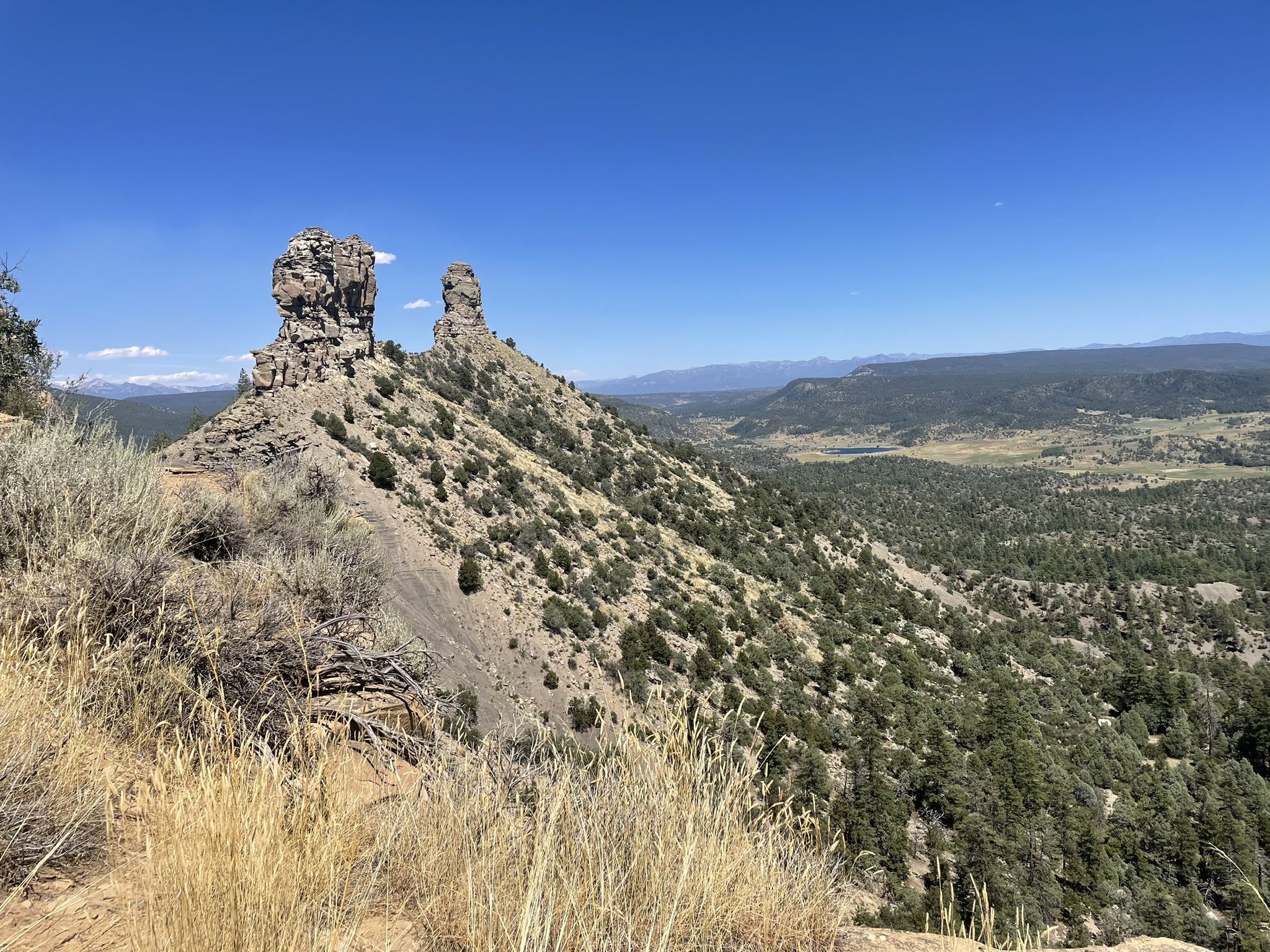Two unique rock formations on a sunny, brushy hillside with distant mountains. - Great House Pueblo Trail