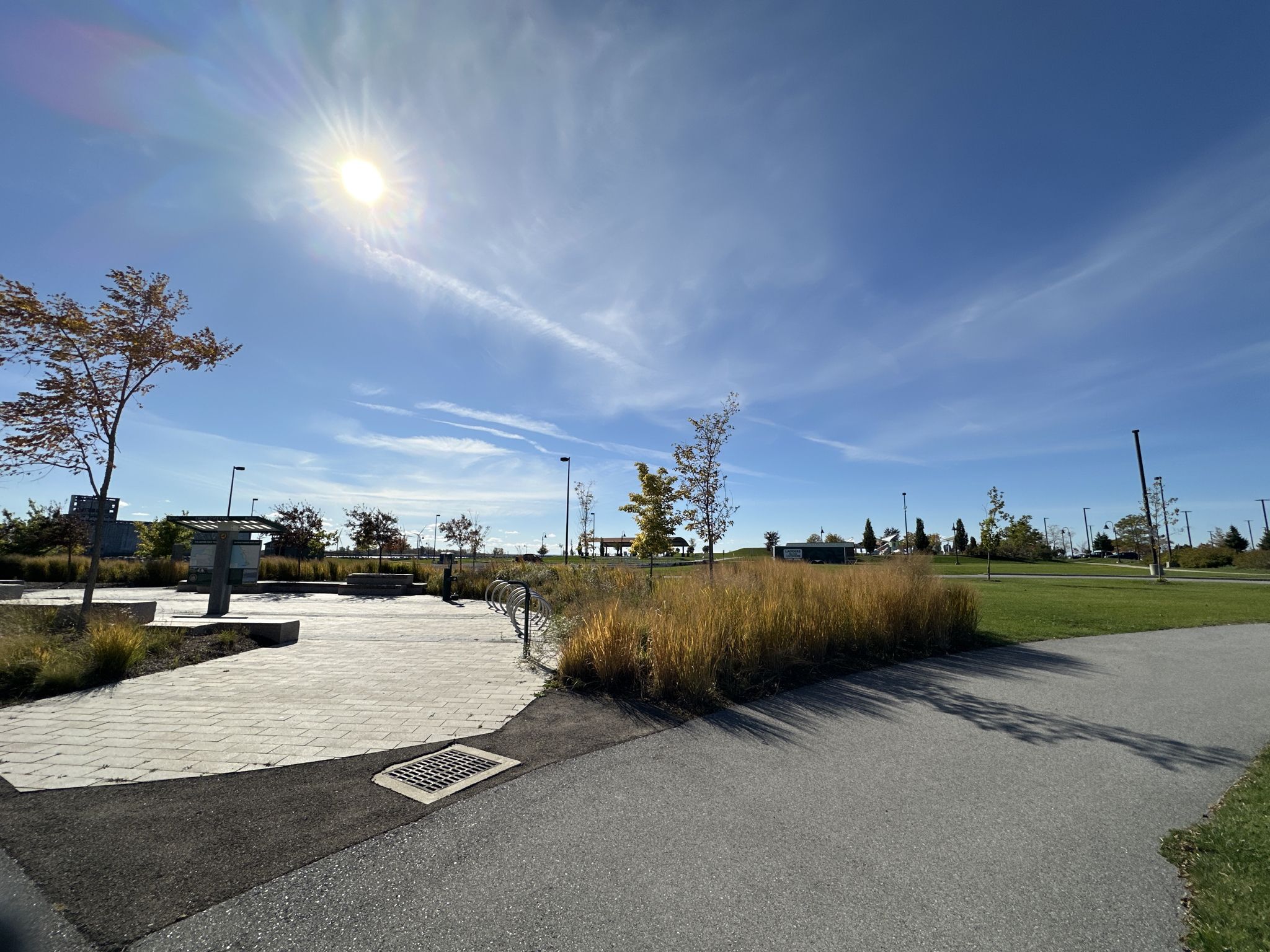 Sunny paved park area with grass, sparse trees, and bright sky. - Fuhrmann Boulevard Sidepath