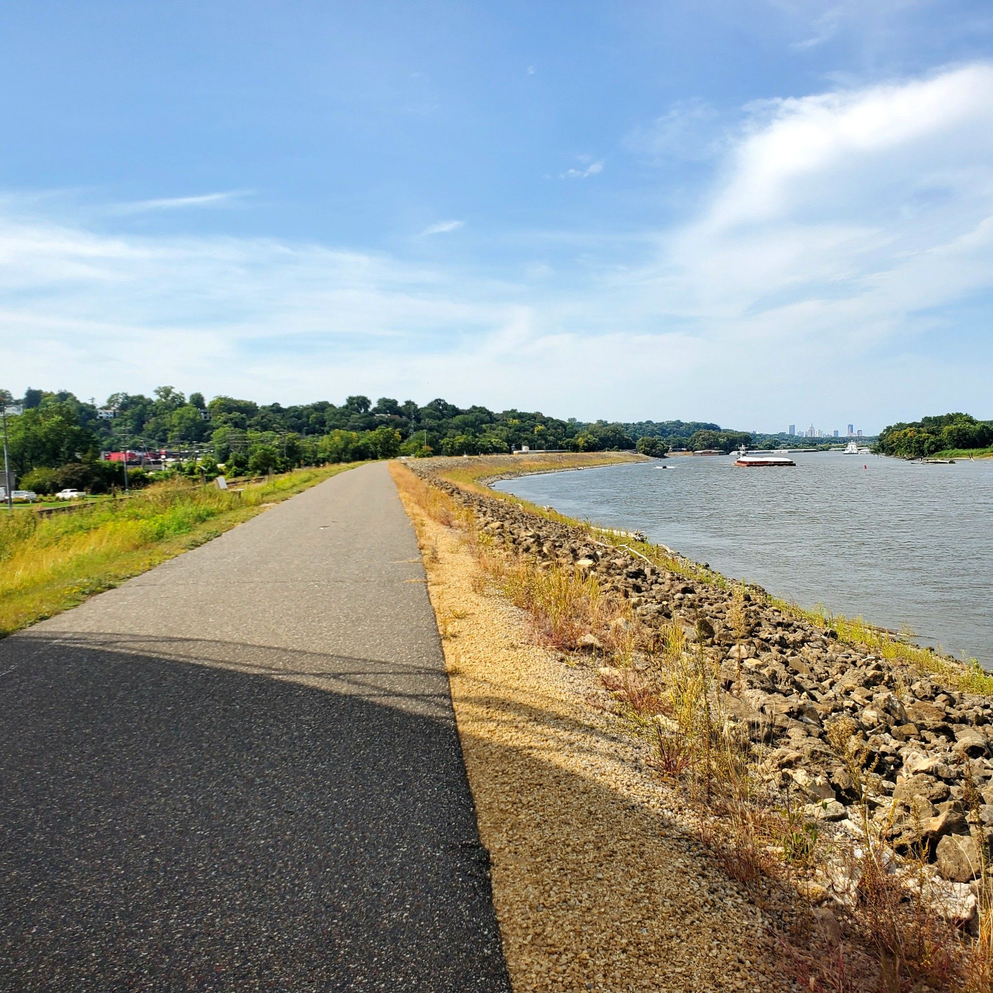 Paved path alongside a wide river with distant boats and green trees. - Mississippi River Greenway