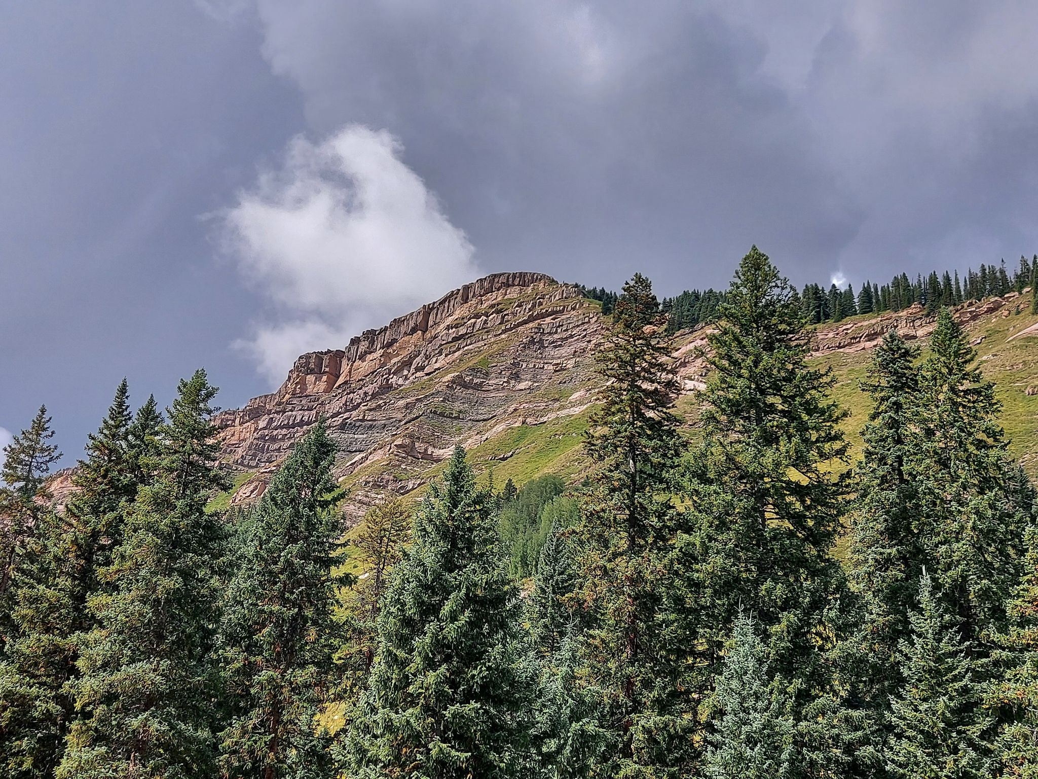 Tall pine trees frame a layered mountain cliff under a dramatic cloudy sky. - Coal Bank Pass – Coal Bank Pass loop from Haviland Lake SWA