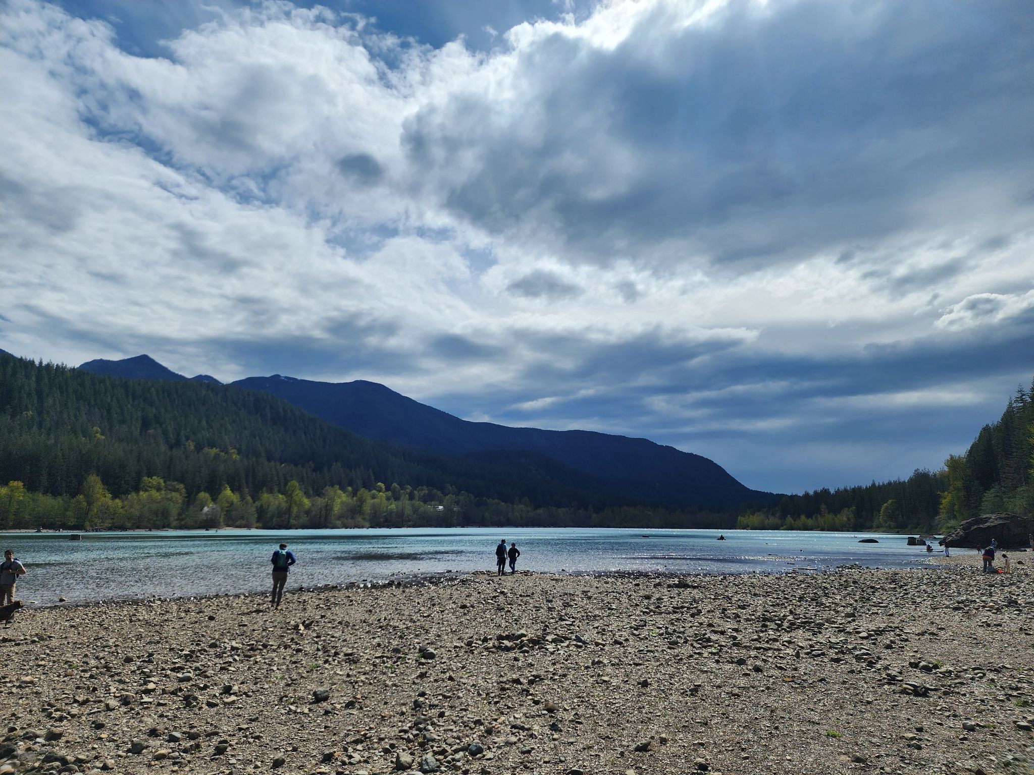 People on a wide rocky beach by a calm lake with forested mountains. - Rattlesnake Ledge Trail
