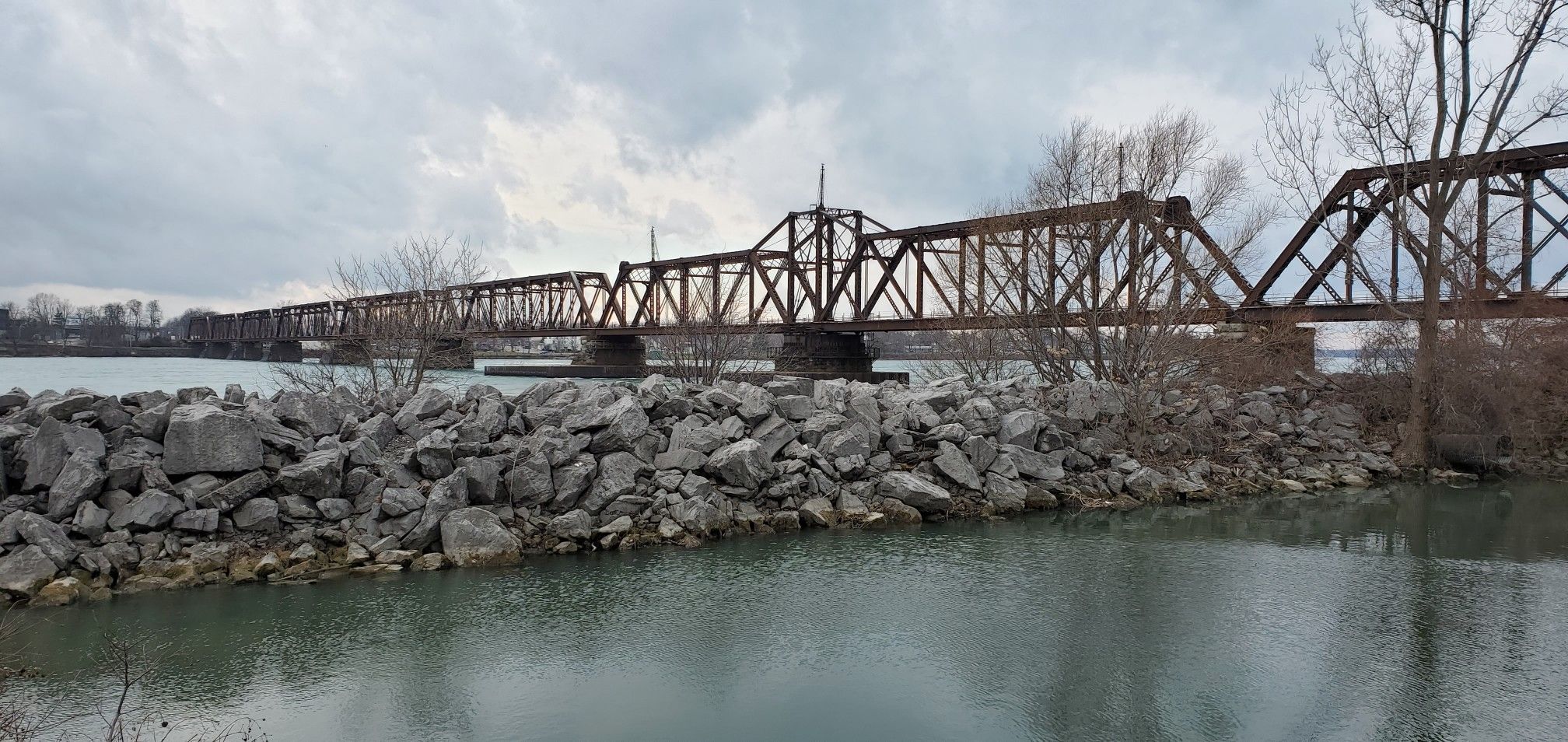 A long, rusty truss bridge spans over water with a rocky shore under a cloudy sky. - Aqua Lane Park – Grand Island Bridges loop from Tonawanda