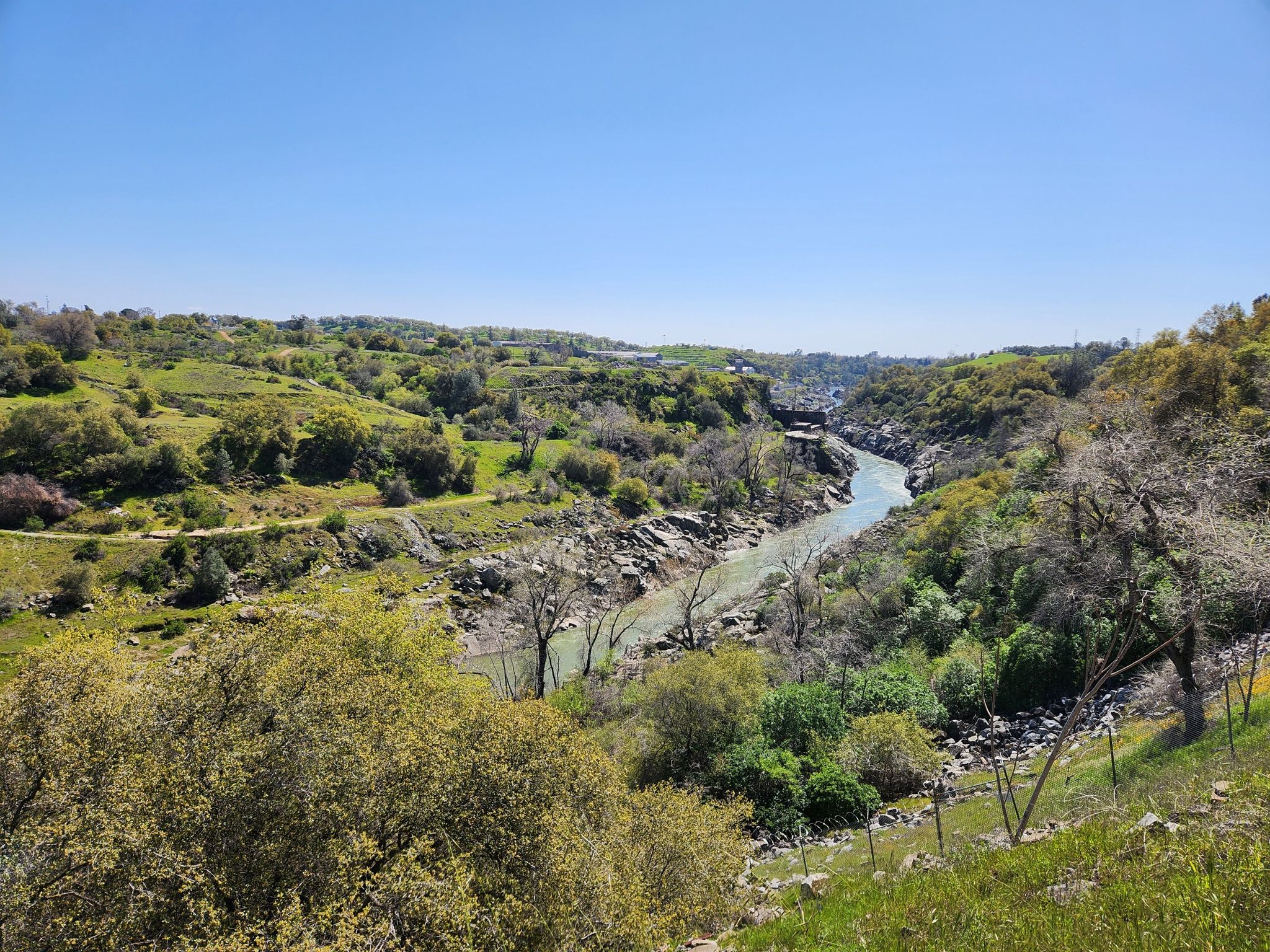 A winding river flows through green, rocky hills under a clear blue sky. - Folsom Historic Truss Bridge – Lake Natoma loop from Iron Point