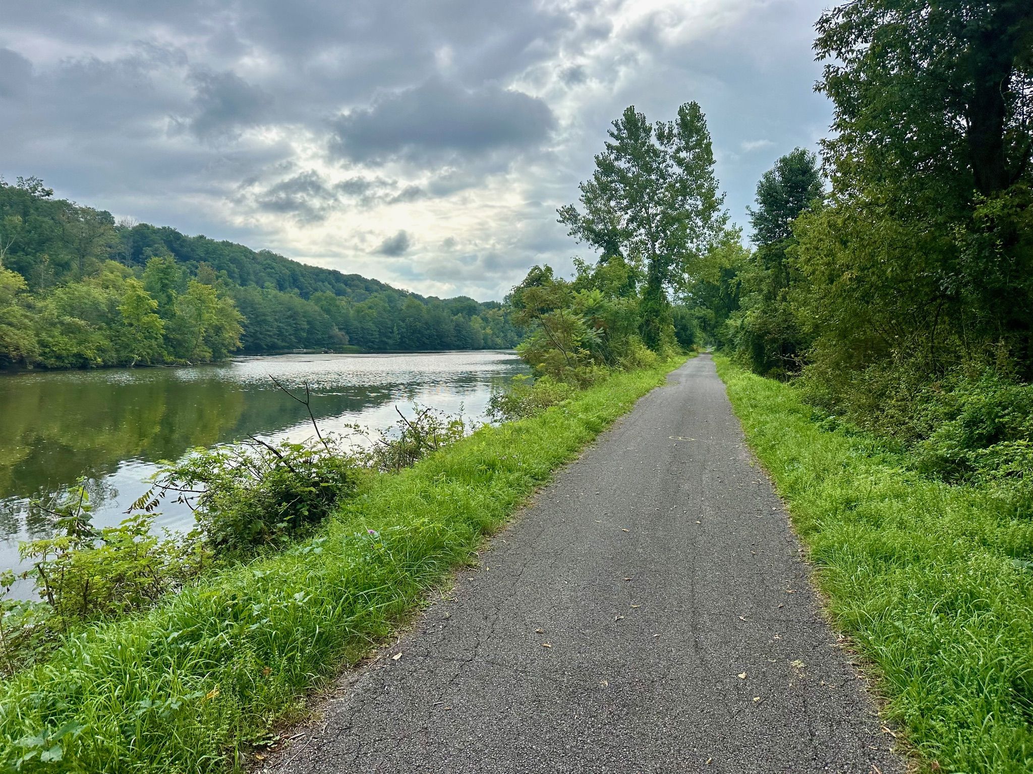 Paved path alongside a calm river with lush green trees under a cloudy sky. - Erie Canalway Trail (Rome to Utica)