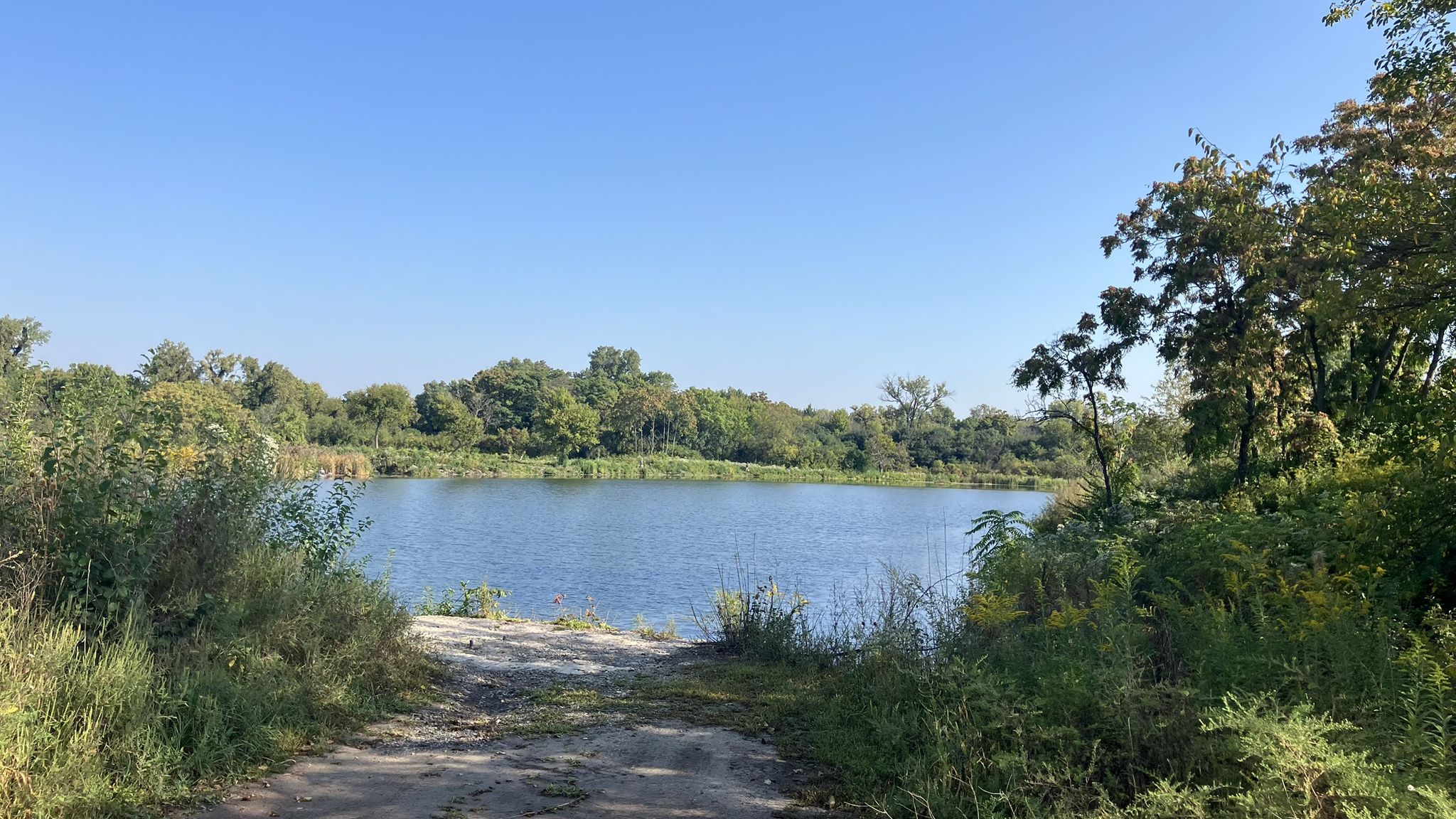 A dirt path leads to a calm lake surrounded by green trees under blue sky. - Heritage Quarries Recreation Area loop