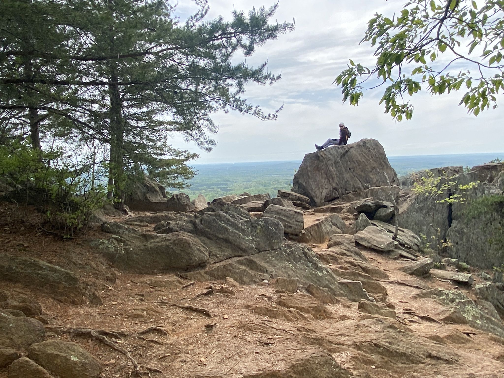 Person sitting on a large rock, overlooking a vast forested landscape under a cloudy sky. - Rocktop and Backside Loop