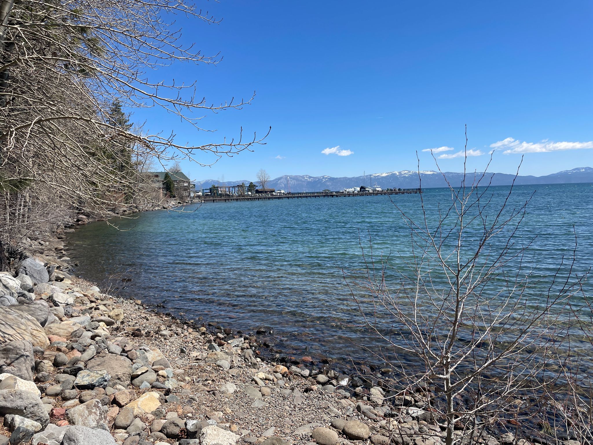 Rocky shoreline, clear blue water, distant mountains, and a pier on a sunny day. - Dollar Point to Tahoe City