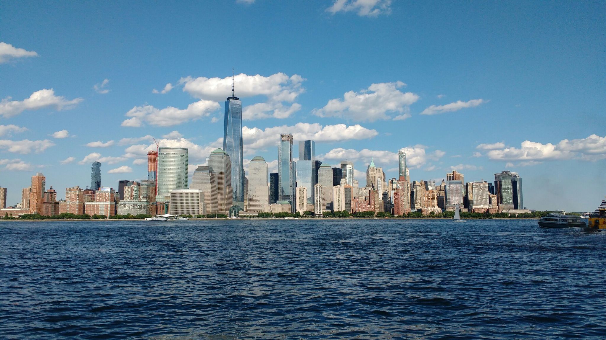 City skyline across water with a boat, under a bright, sunny sky. - Jersey City 9/11 Memorial – View of the Manhattan Skyline loop from Harsimus Cove