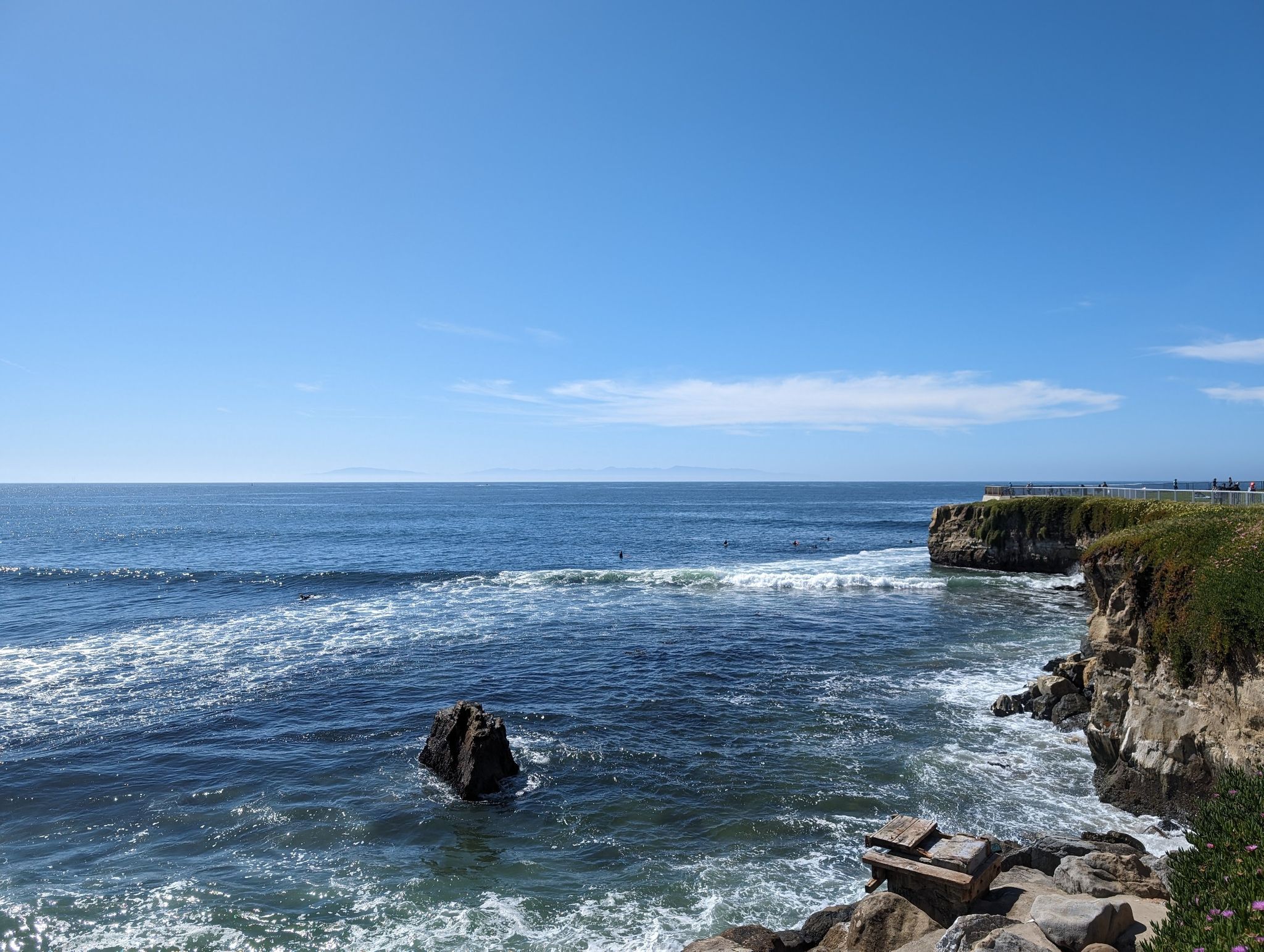 Sunny ocean view with distant surfers, rocky coastline, and clear blue sky. - West Cliff Cycleway – Santa Cruz Lighthouse loop from Lighthouse Field State Beach