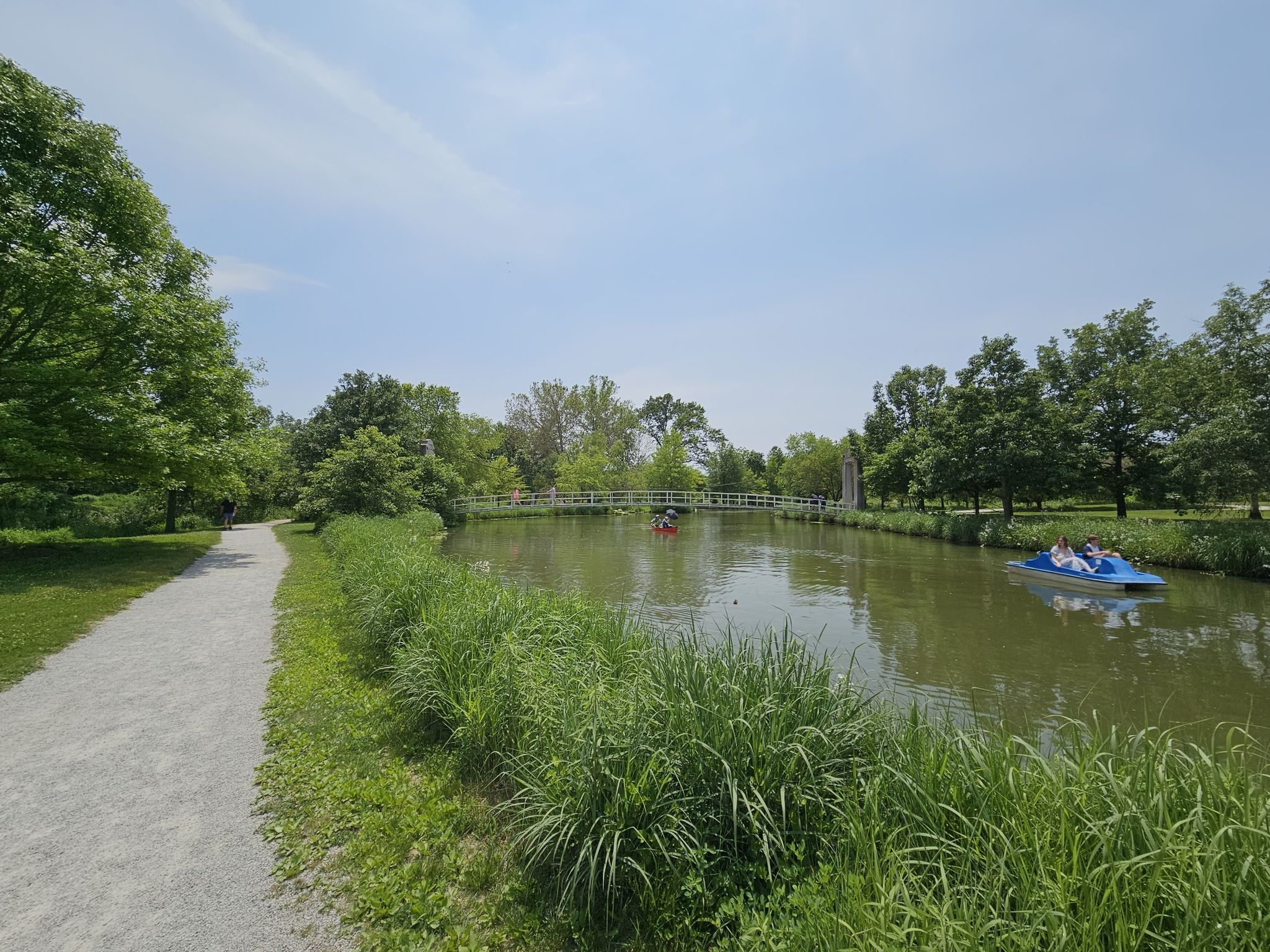 People paddle boating on a pond surrounded by green trees and a path. - Emerson Grand Basin Loop