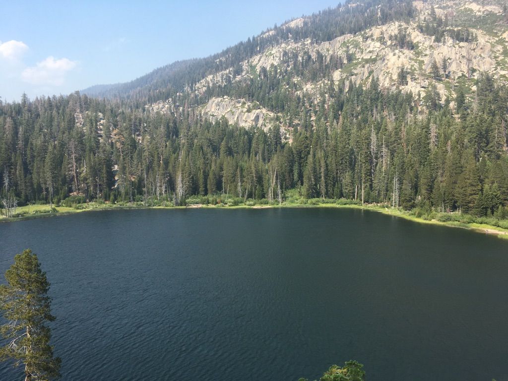 A dark blue lake surrounded by a dense green forest and rocky mountains on a clear day. - Sotcher Lake Loop