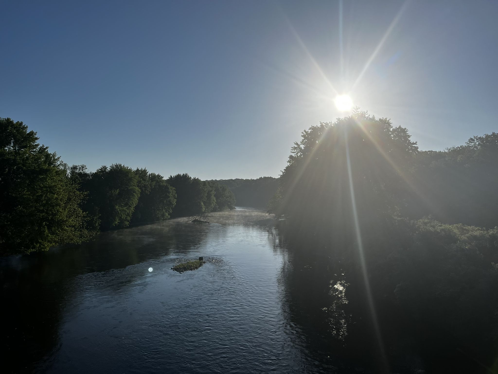 A wide river with green trees on banks, bright sun flare in a clear sky. - Fred Meijer White Pine Trail