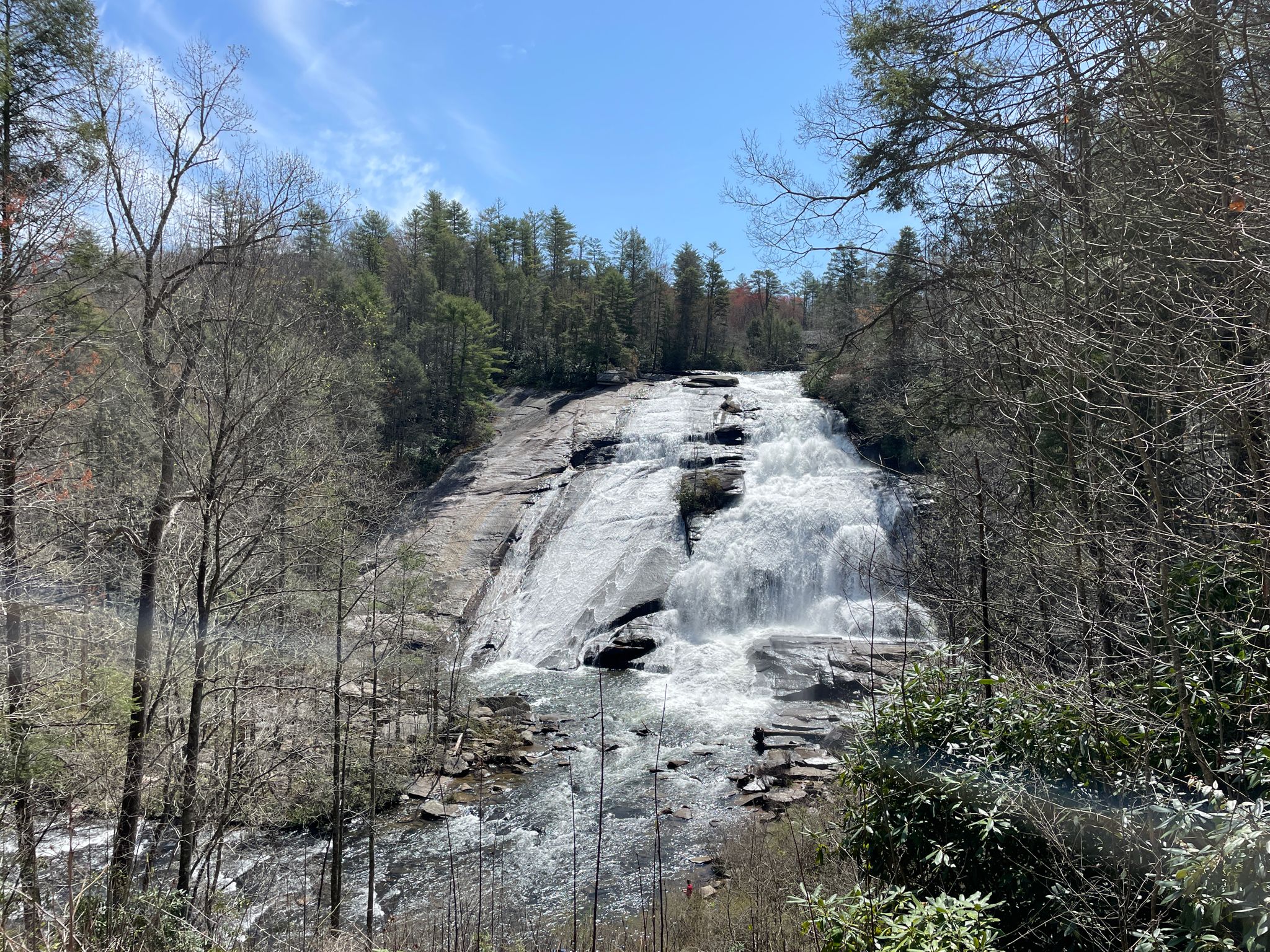 Wide waterfall flows over rock formations, surrounded by trees under blue sky. - High Falls Loop