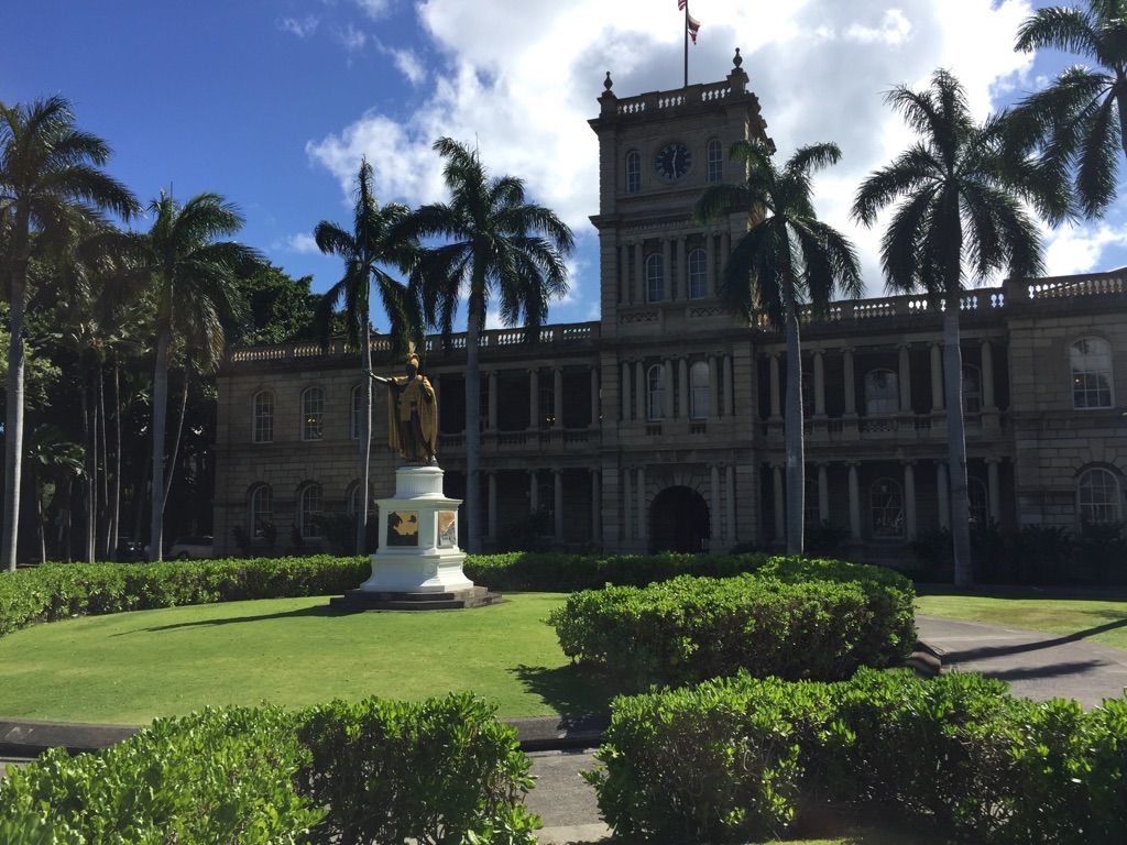 Historic building with clock tower, statue, palm trees, and green lawn. - Hawaii State Capitol and Palace Grounds