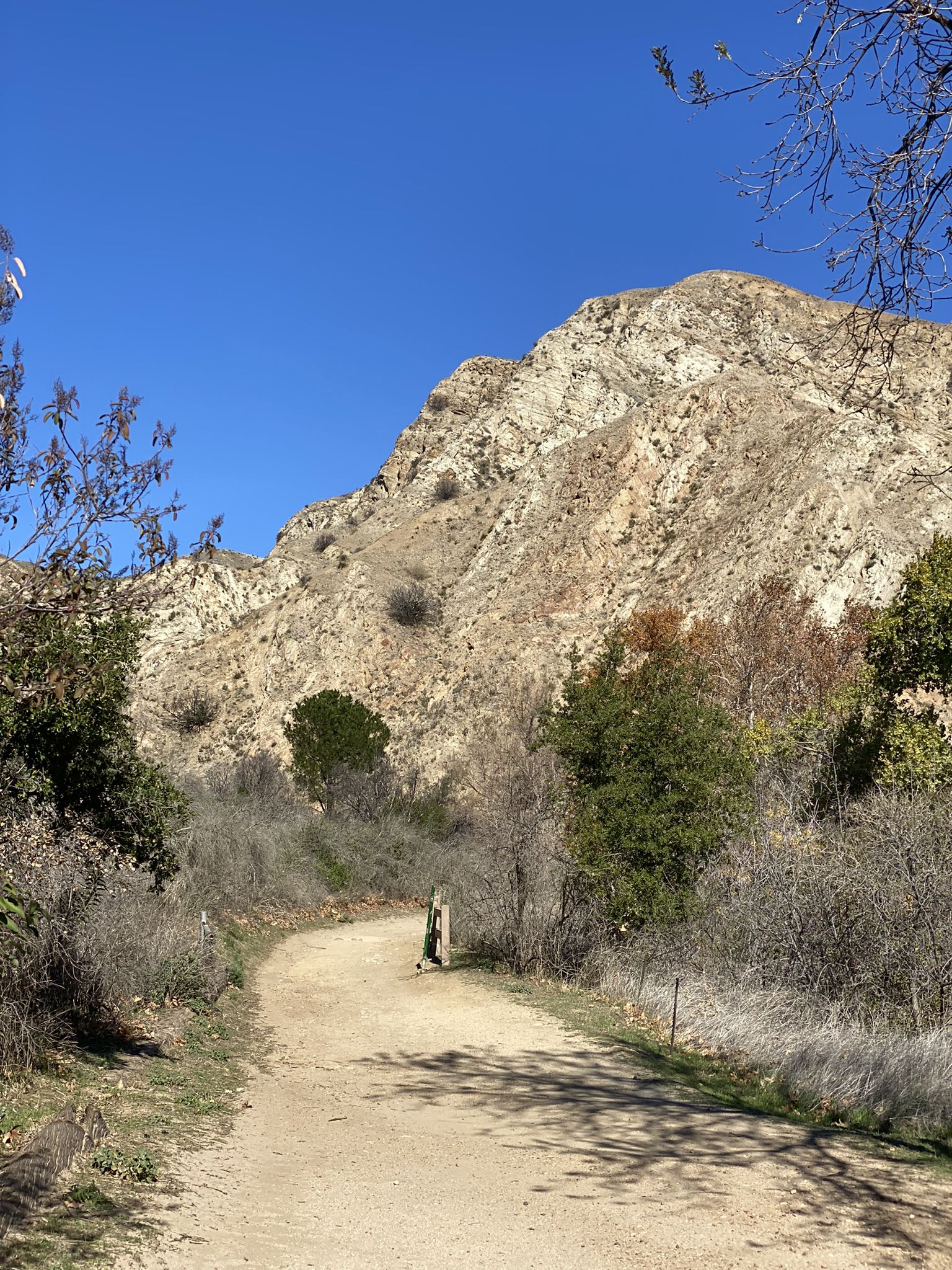 A dirt road leads towards a large, rocky mountain under a clear blue sky. - Entrance to O'Melveny Park – View of Mission Point loop from O'Melveny Park