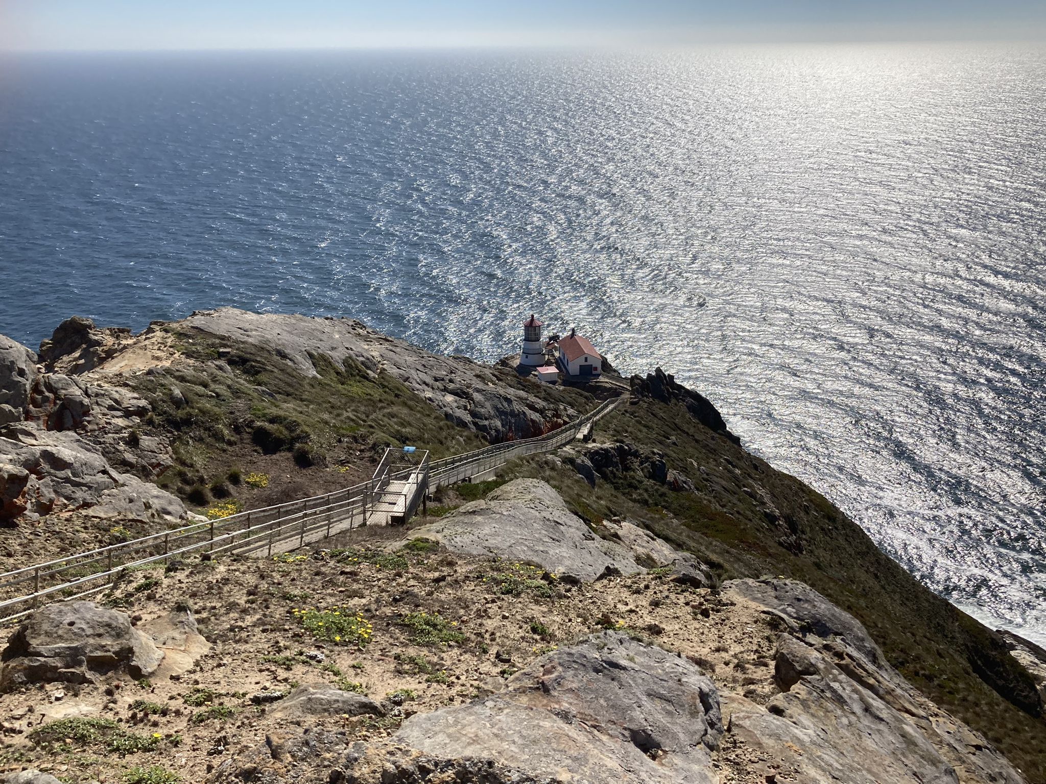 Fenced walkway leading to a lighthouse on a rocky cliff, bright sun. - Point Reyes Lighthouse Trail