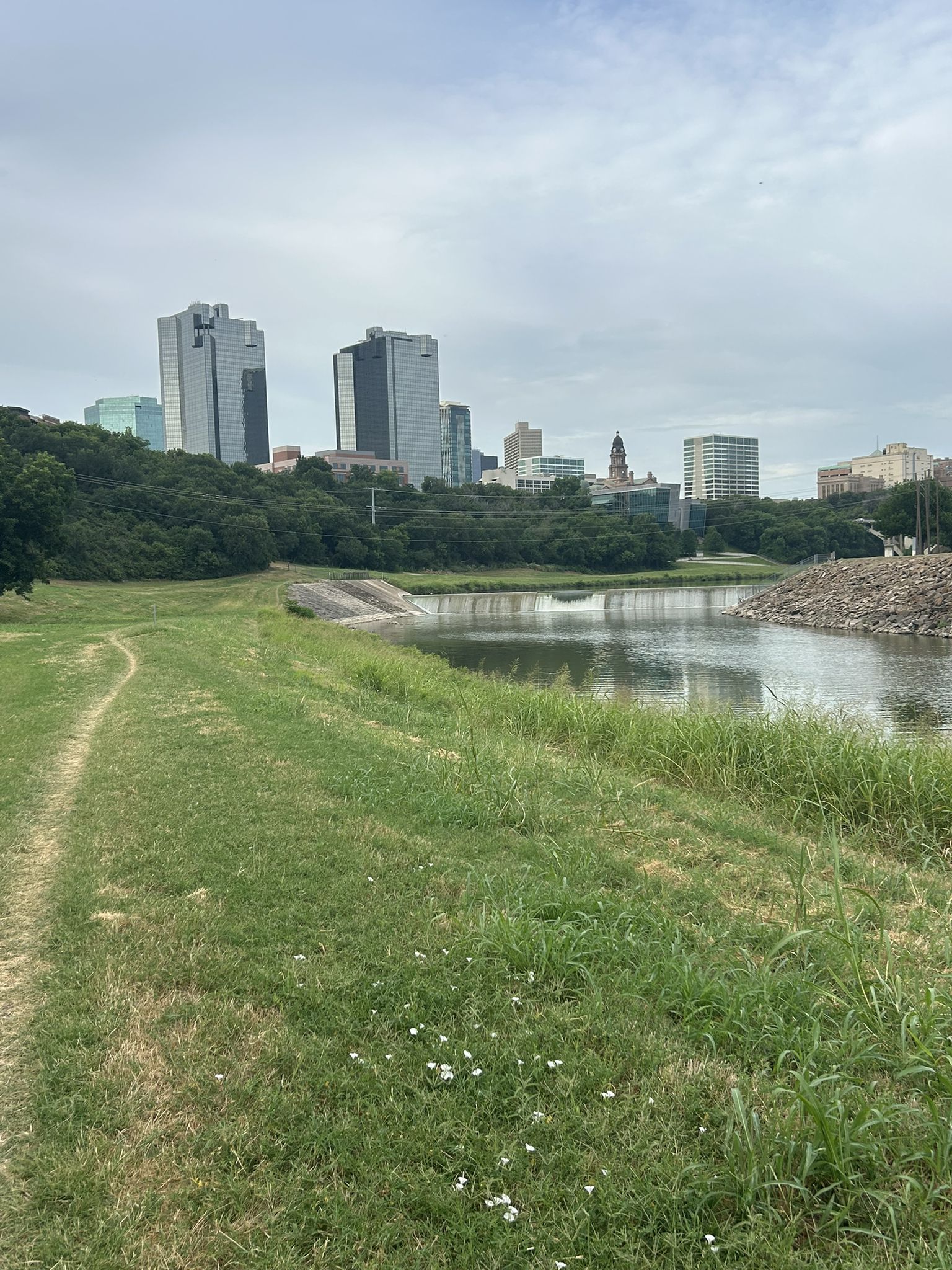 Overcast view of a river, grassy bank, and distant city skyline. - Trinity Trails: West Fork Loop