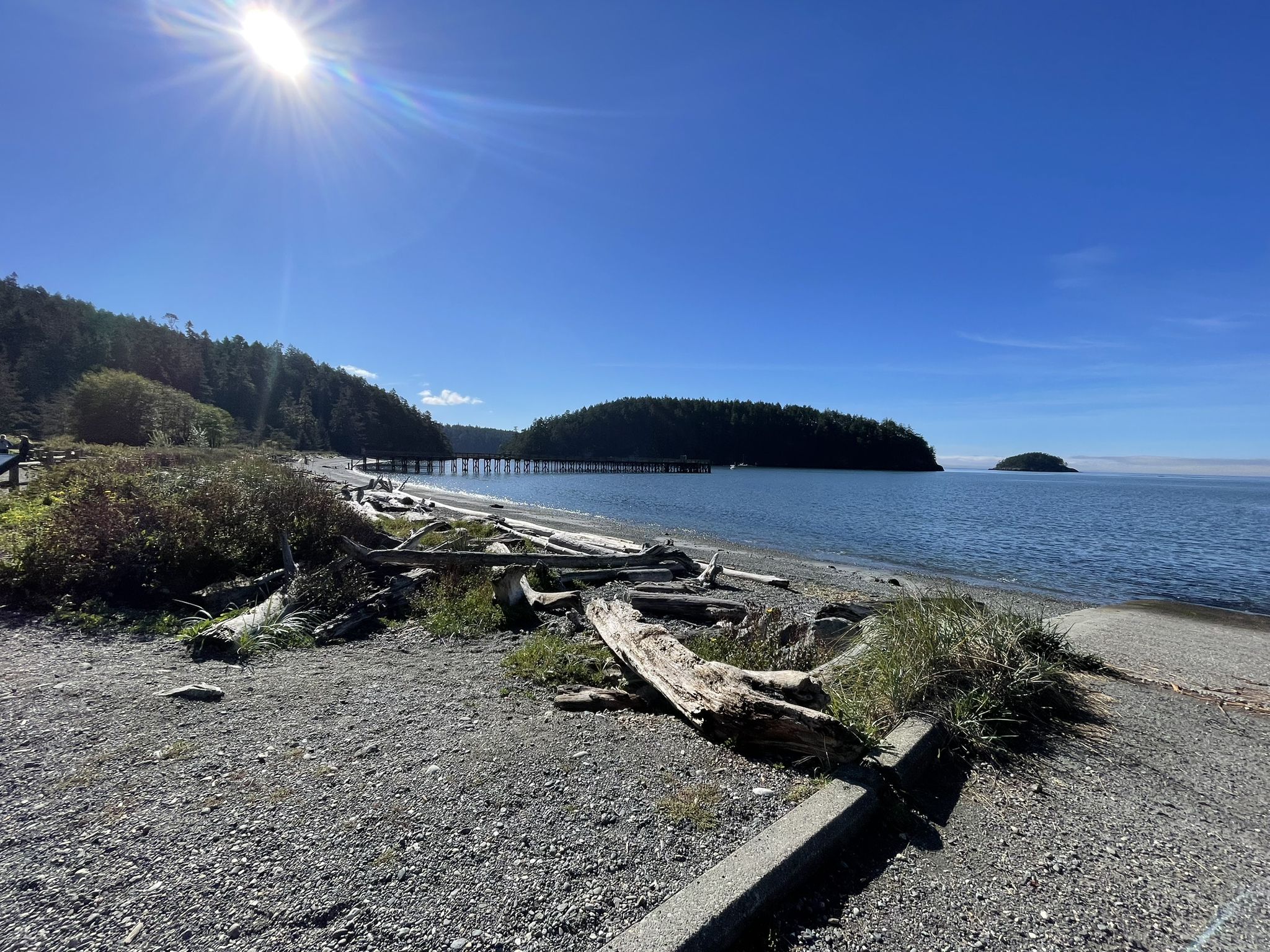 Wide pebbly beach with driftwood, calm blue water, and a bright sunny sky. - Pass Lake, Ginnett Hill, and John Tursi Trail Loop