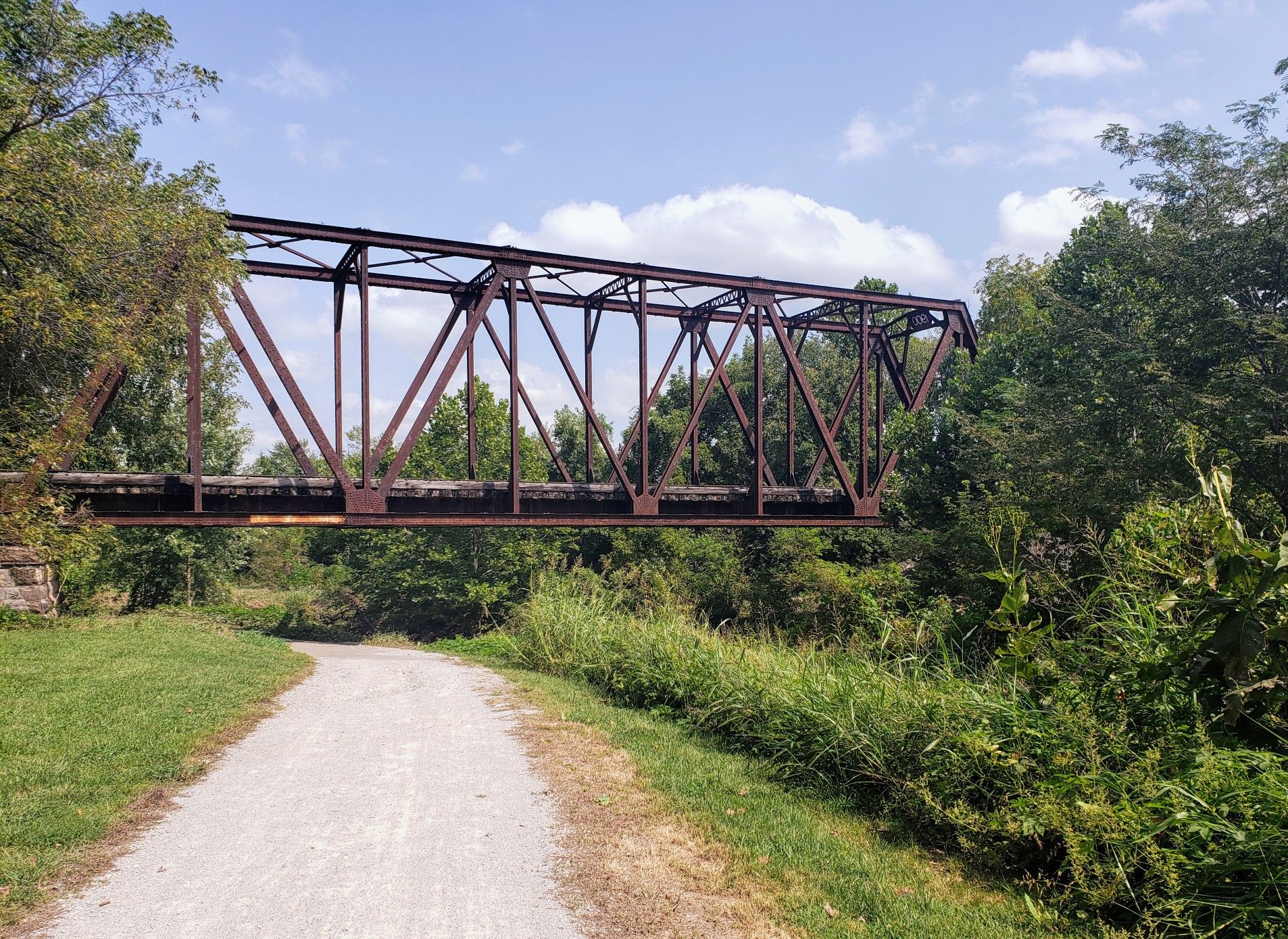 An old metal truss bridge spans over a path, surrounded by green foliage. - Little Blue Trace Trail – Little Blue Trail Park loop from Independence