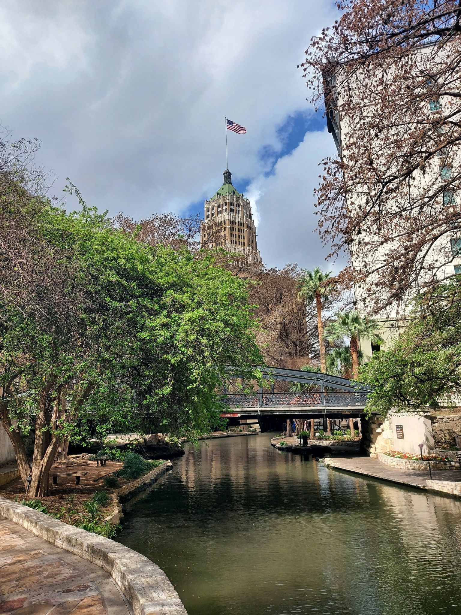 River with bridge, tall building, and lush trees under a cloudy sky. - San Antonio River Walk Loop