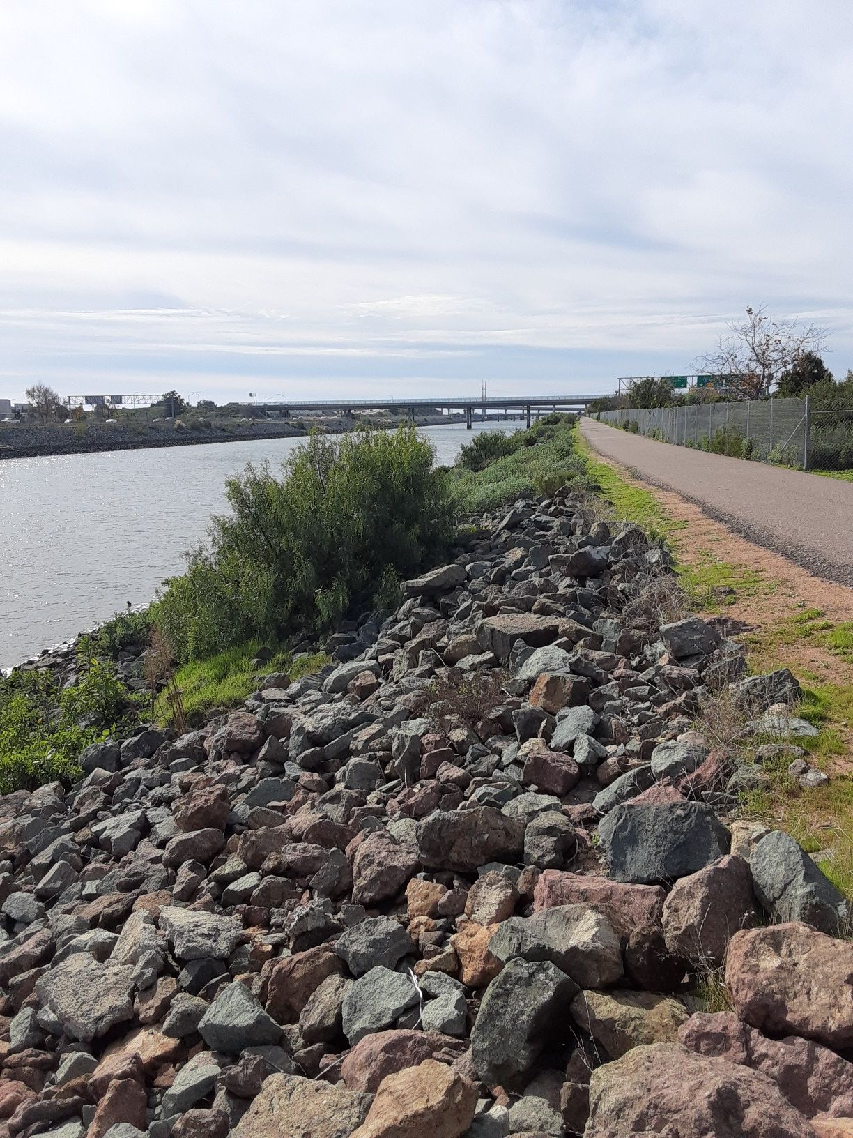 Paved path alongside a rocky riverbank under a cloudy sky. - Bayshore Bikeway – Sweetwater River loop from Southwestern College