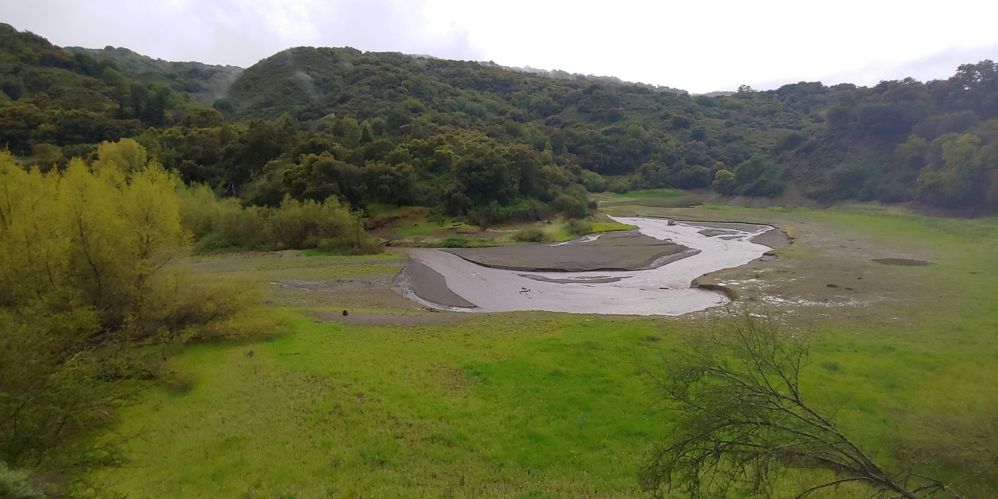Overcast view of a muddy stream flowing through a wide green valley. - Stevens Creek County Park – Stevens Creek Dam loop from Picchetti Ranch Open Space Preserve