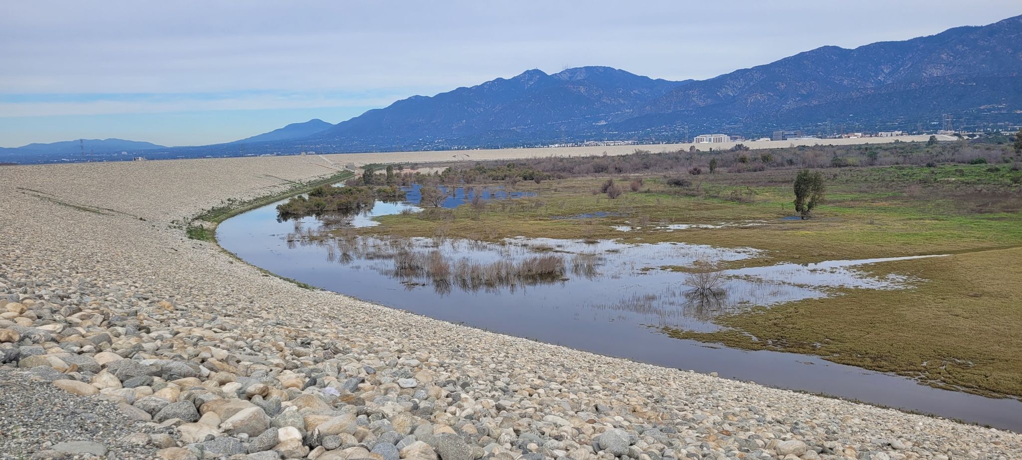 A rocky embankment next to a winding river and green wetlands. - Santa Fe Flood Control Basin loop from Irwindale