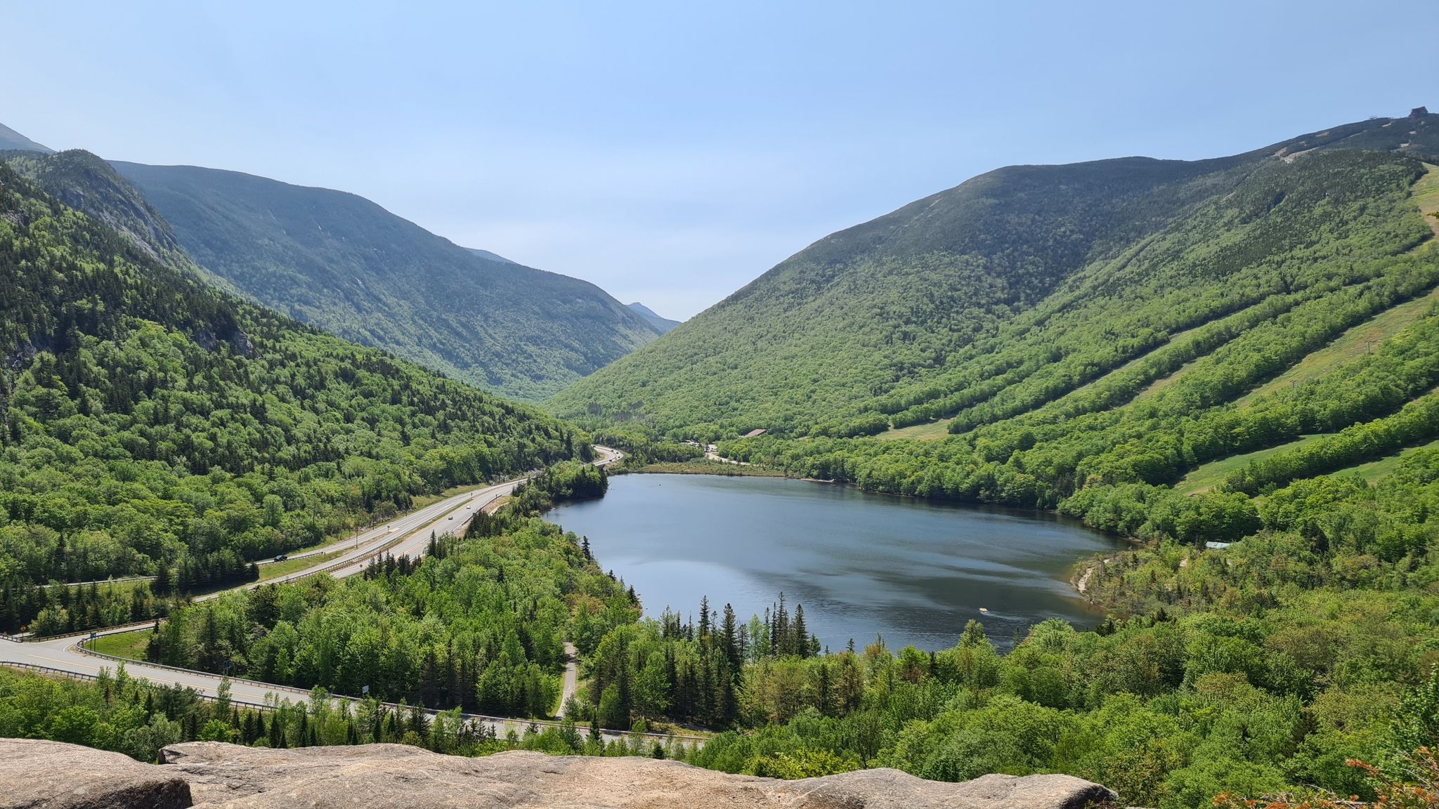 A serene lake nestled between green mountains with a highway nearby on a clear day. - Bald Mountain and Artist's Bluff Loop