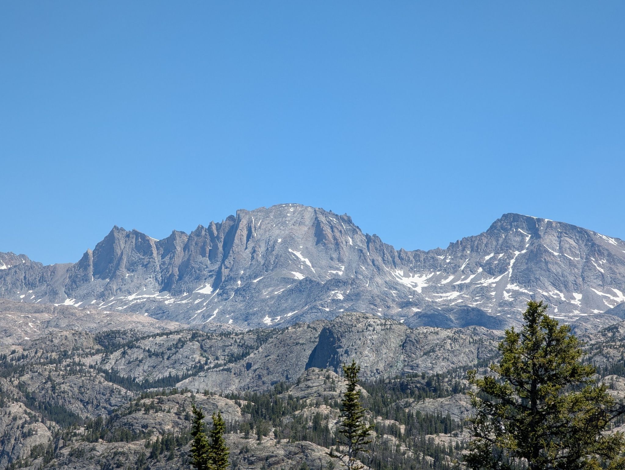 Majestic, snow-dusted mountains rise above a rocky, forested landscape under bright sun. - Miller Lake and Upper Sweeney Lake Loop