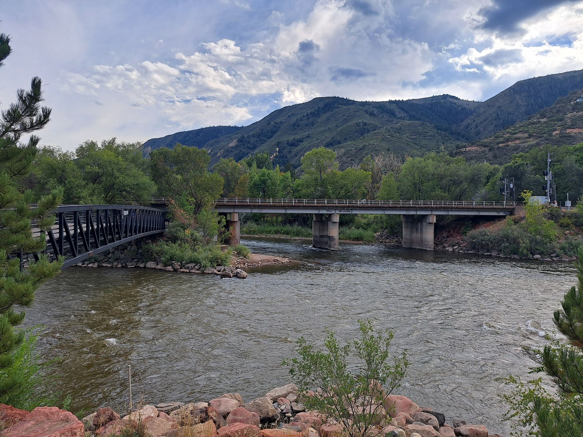 Two bridges span a river, surrounded by green trees and distant mountains. - View of Roaring Fork River – Two Rivers Park loop from Sopris Elementary School