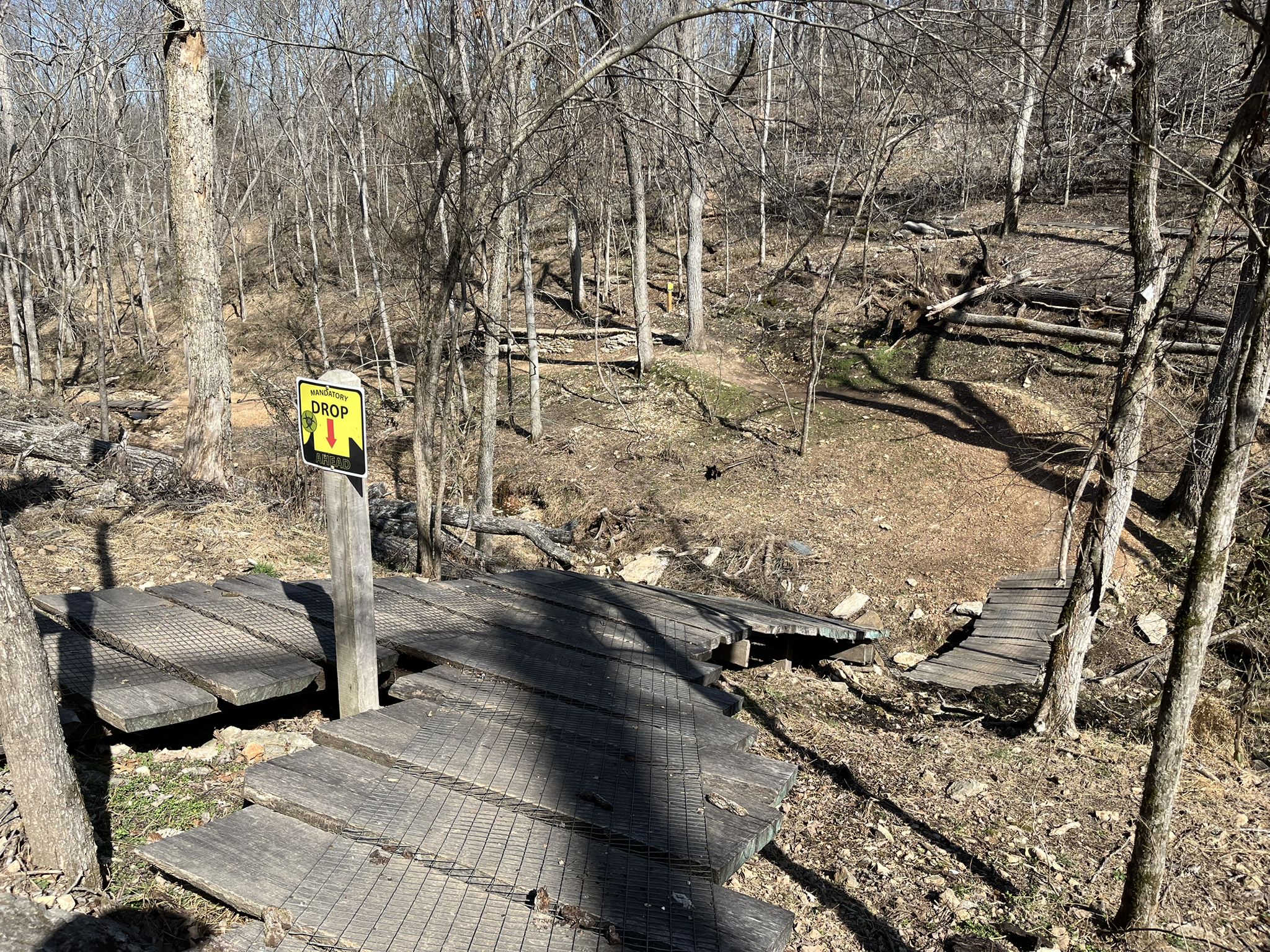 Wooden bike drop feature with a warning sign in a sunny, bare forest. - Slaughter Pen Trails – Bentonville City Square loop from Bentonville Municipal Airport