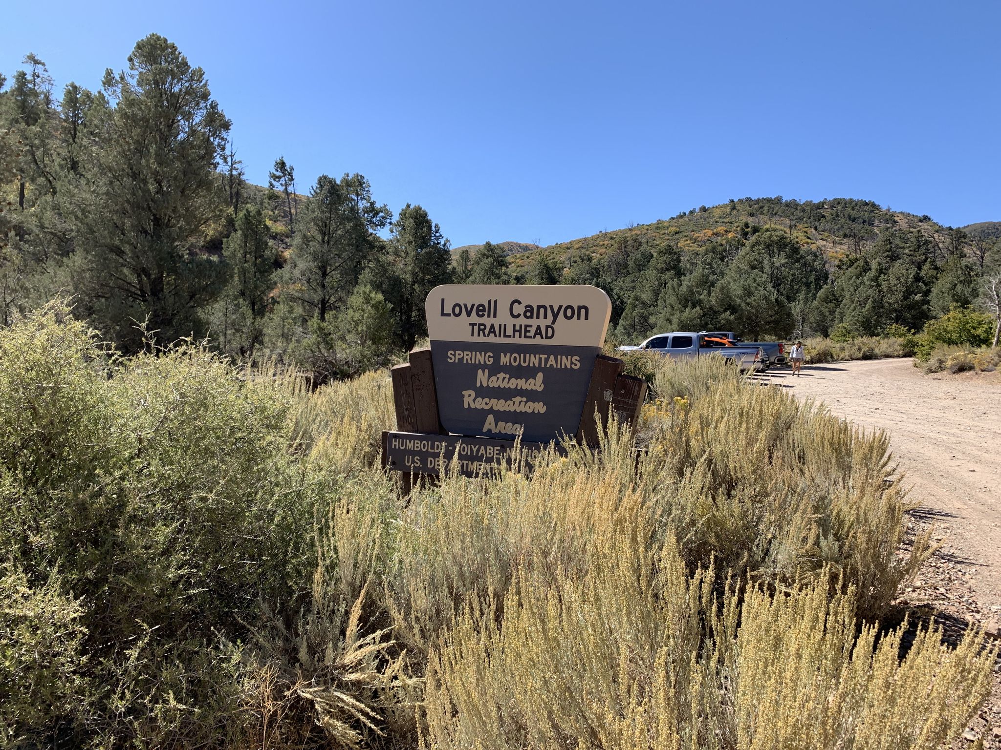 A wooden sign stands amidst tall brush on a sunny day, with a truck and hills. - Lovell Canyon and Lovell Summit Road OHV Route