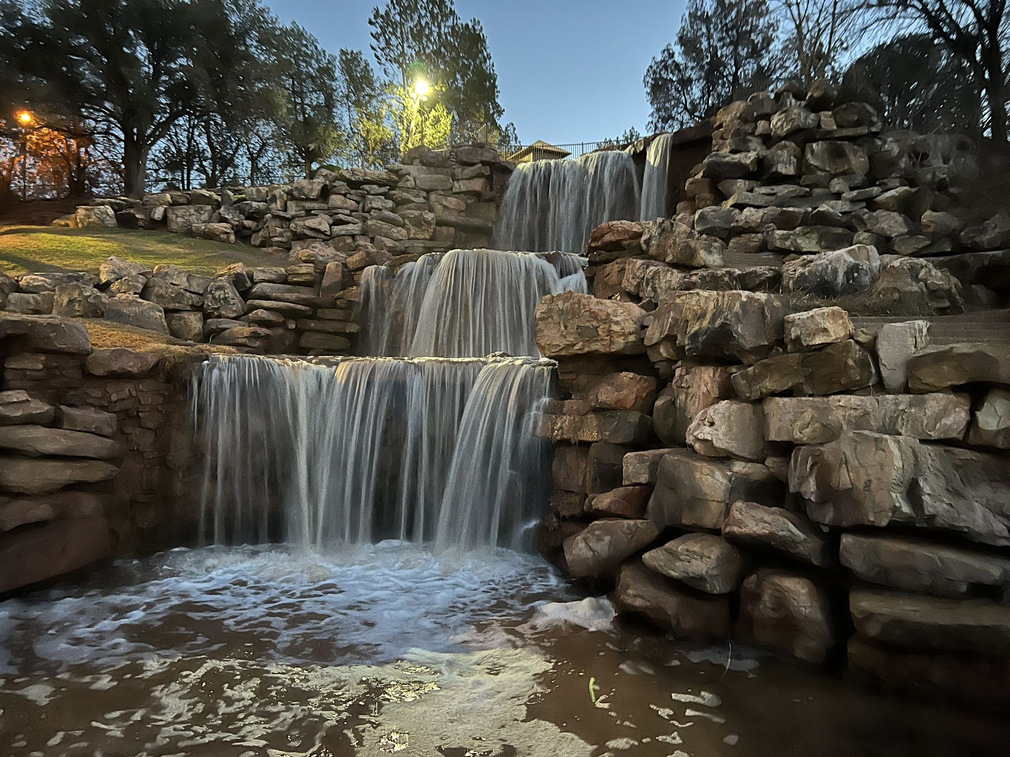 Multi-tiered rock waterfall illuminated by artificial lights at dusk, with trees in background. - Lucy Park Circle Trail