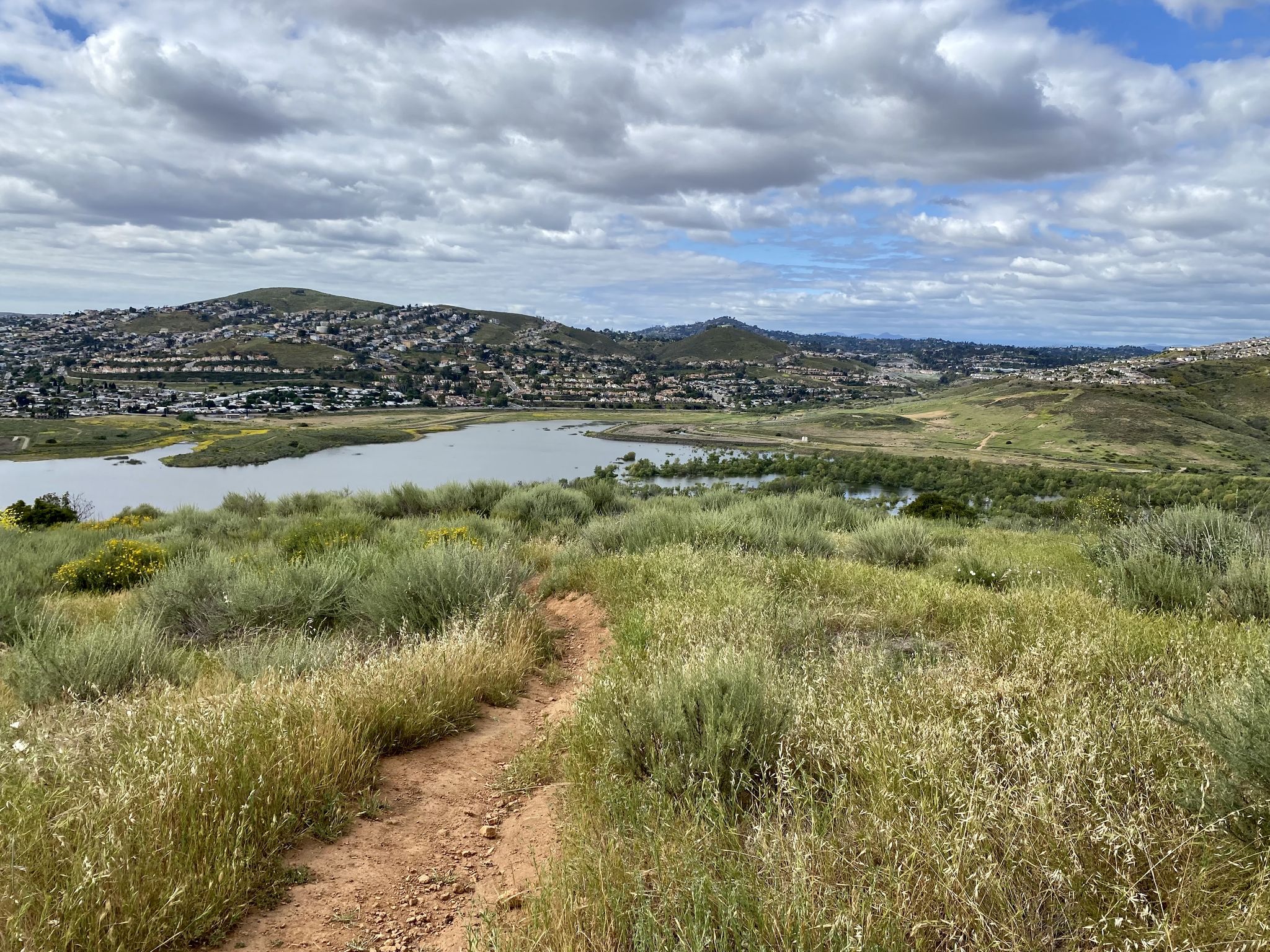 Dirt path through green hills overlooking a lake and distant town. - Tiki Hut – Sweet Water River Trail loop from La Presa