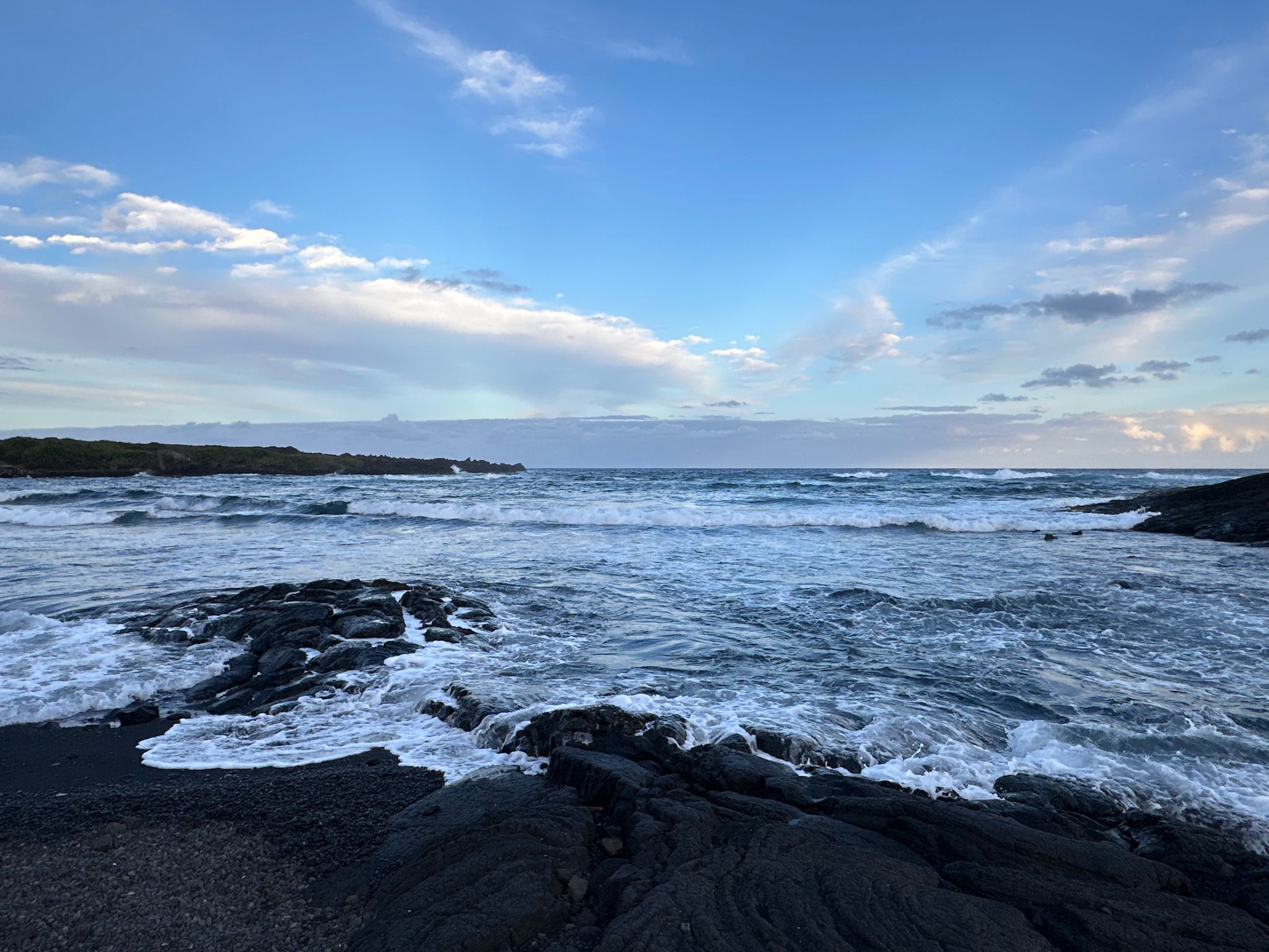 Ocean waves crash on dark volcanic rocks under a partly cloudy blue sky. - Ninole Loop Road