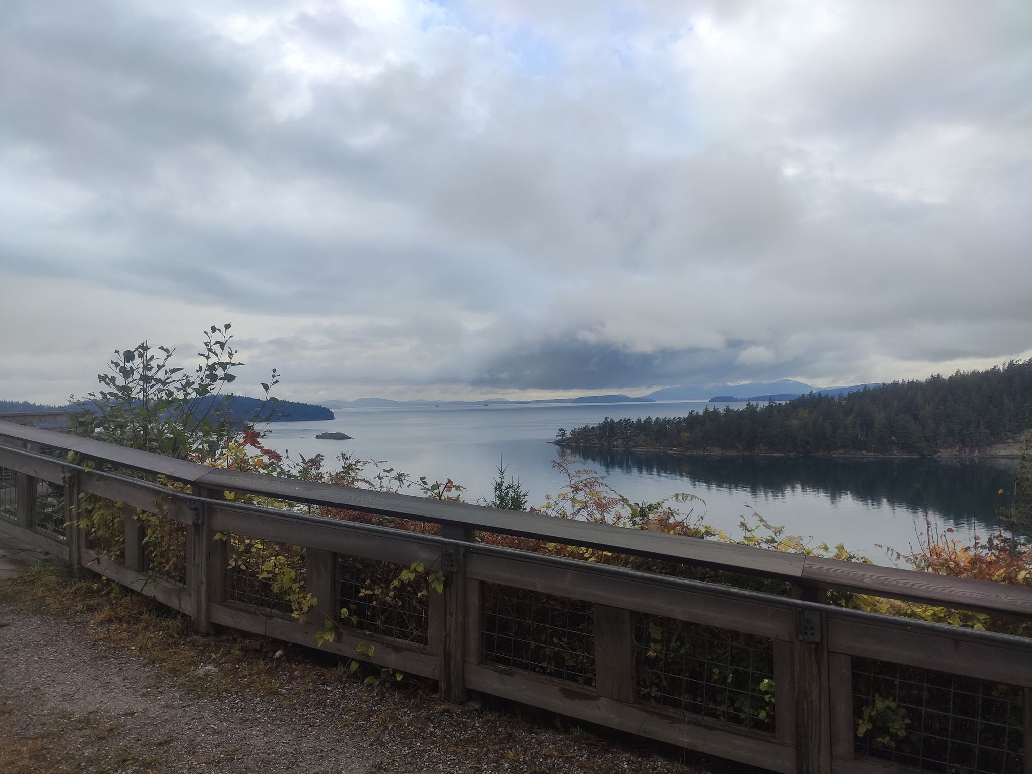 Overcast view of a calm bay with forested islands and a wooden fence. - Interurban Trail Loop