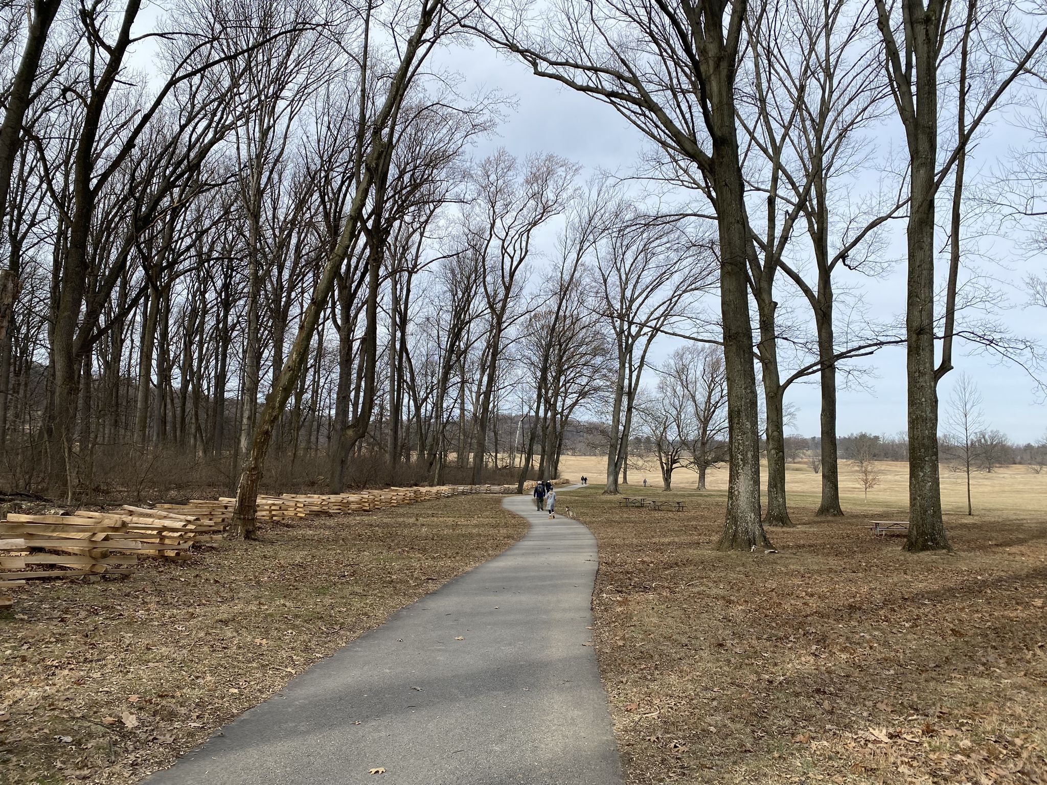Paved path, bare trees, wooden fence, people walking, cloudy sky. - Statue of General Von Steuben – Varnum's Quarters loop from Valley Forge National Historical Park