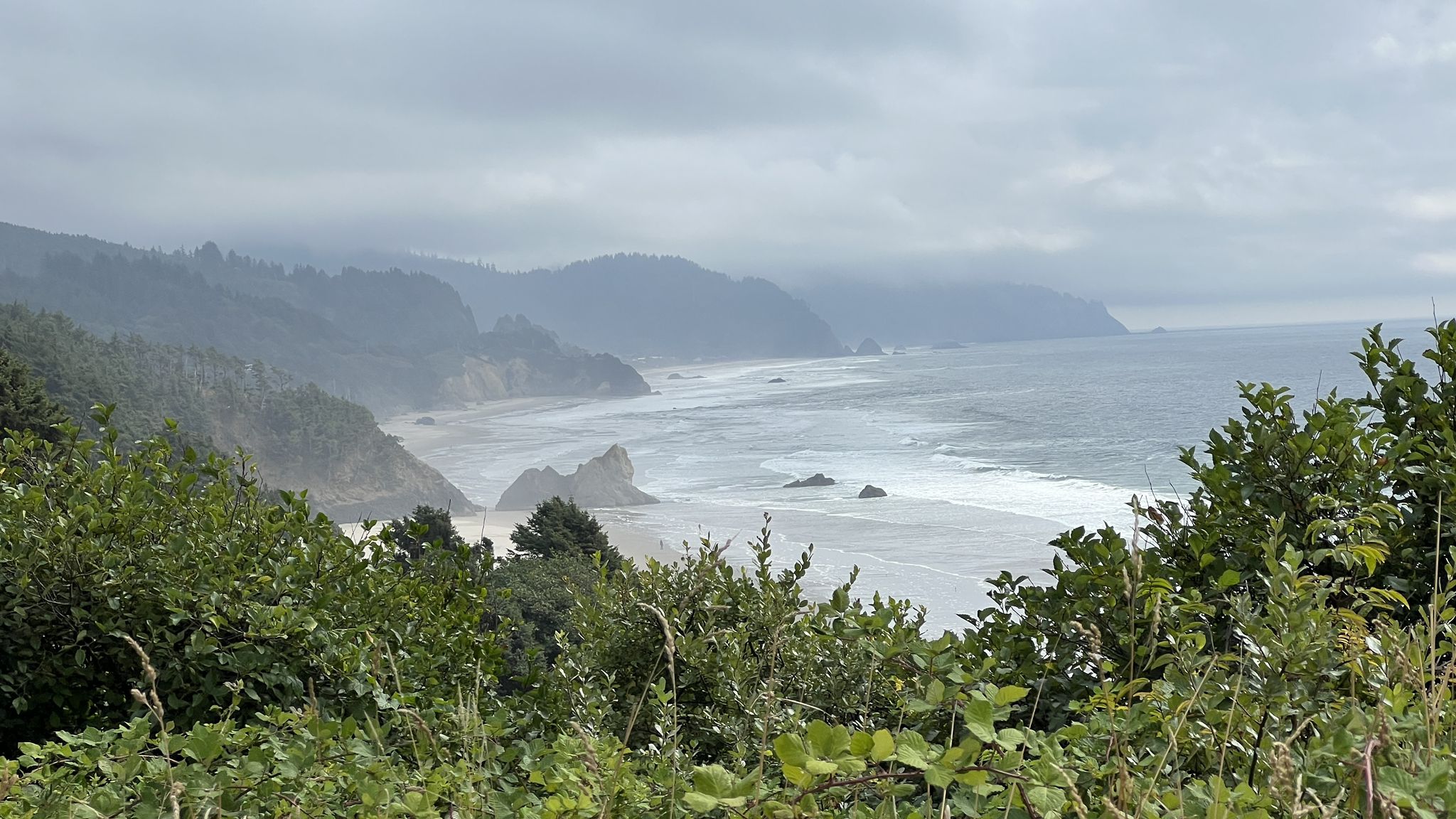 Overcast view of a rugged coastline, sandy beach, and ocean with breaking waves. - Lewis and Clark Road – Haystack Rock loop from Circle Creek
