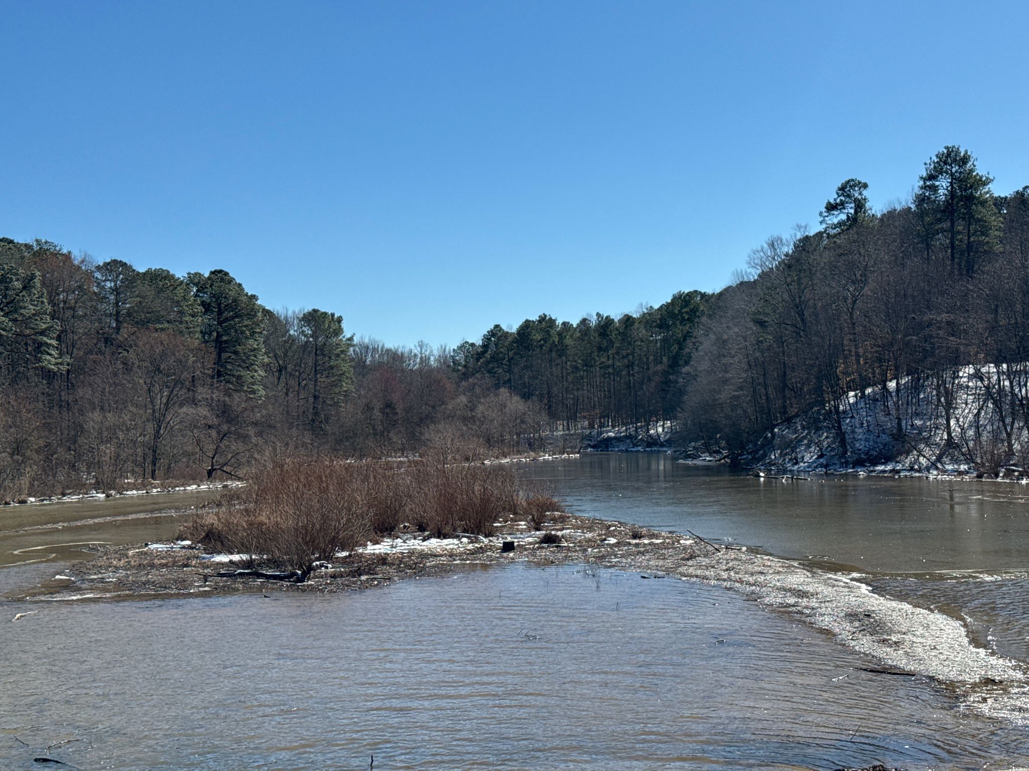 A river flows through a winter landscape with snowy banks and bare trees. - Lake Crabtree Loop Trail