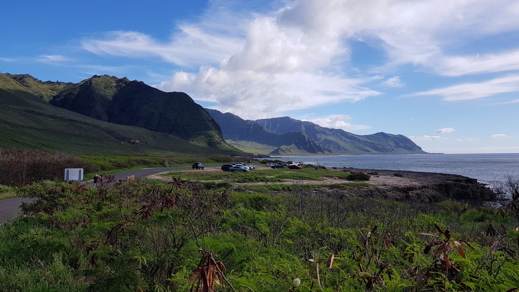 Coastal landscape with green mountains, ocean, and parked cars under a cloudy sky. - Heart-Shaped Rock Hole – Sea arch at Kaʻena Point loop from Kaena Point Satellite Tracking Station