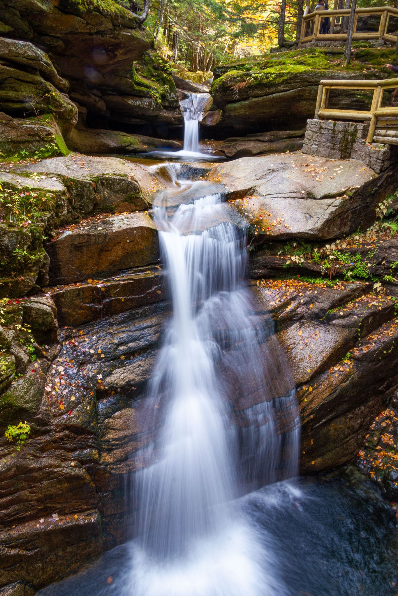 Multi-tiered waterfall cascades over mossy rocks, with autumn leaves and a wooden railing. - Sabbaday Brook Trail