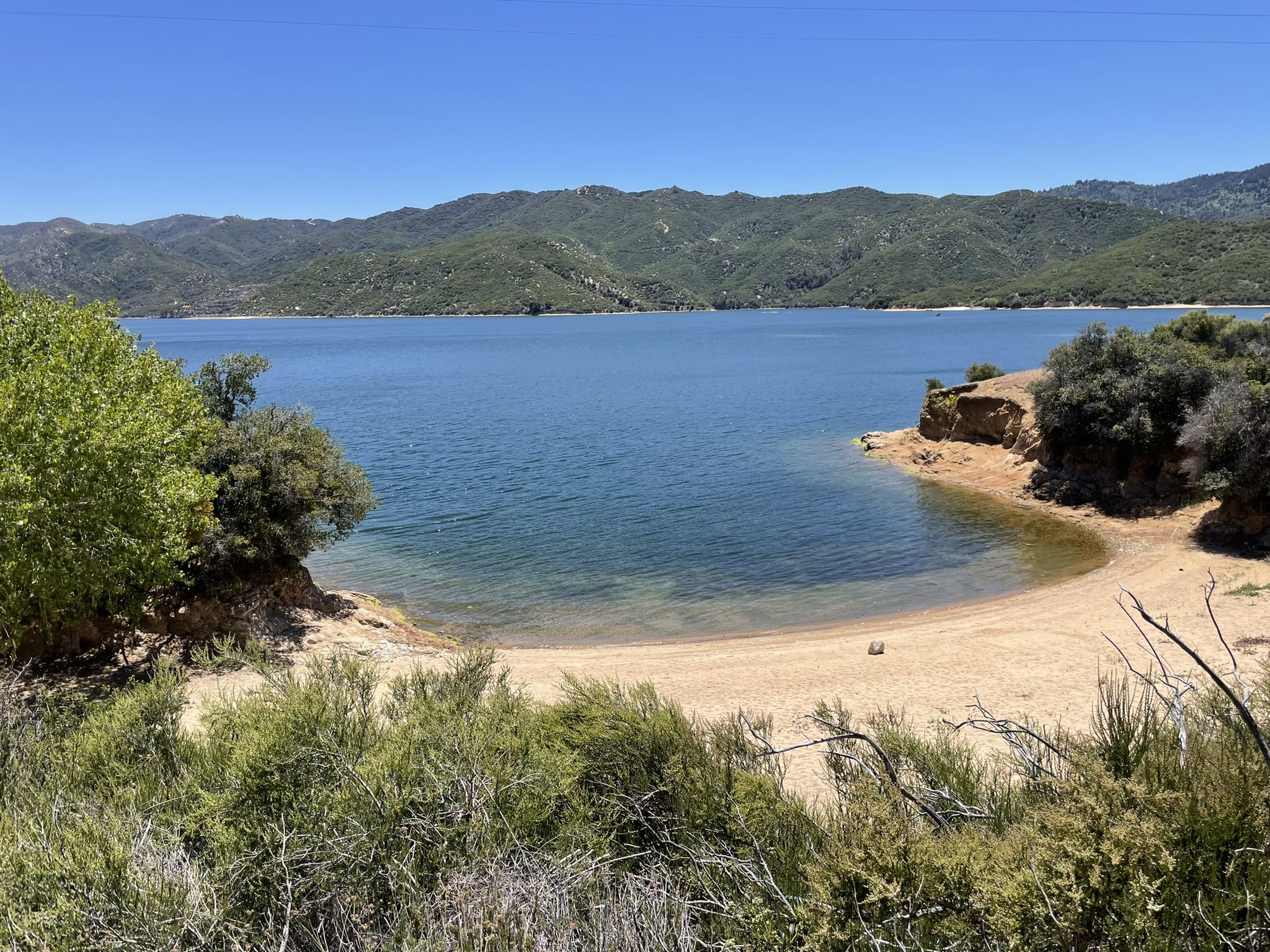 Sunny view of a clear blue lake with a sandy cove and green hills. - Pacific Crest Trail: Silverwood Lake North Shore