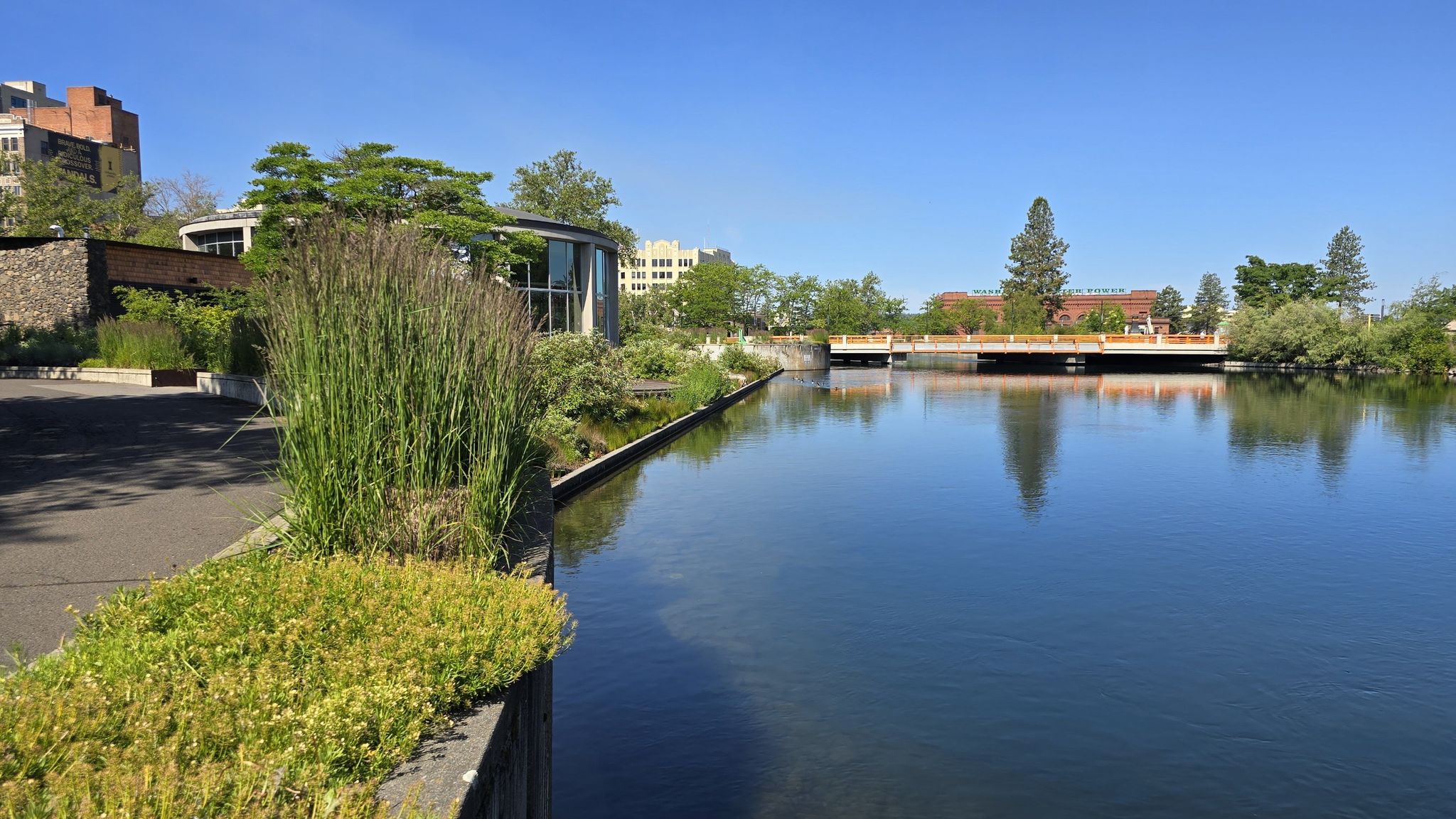 Calm river next to a modern building and green landscaping on a sunny day. - Garbage Goat Sculpture – Riverfront Park loop from Spokane
