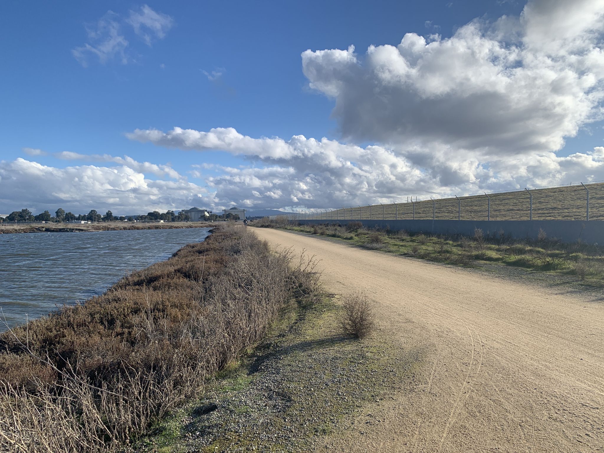 A dirt path runs beside calm water under a bright, cloudy sky. - San Francisco Bay Trail