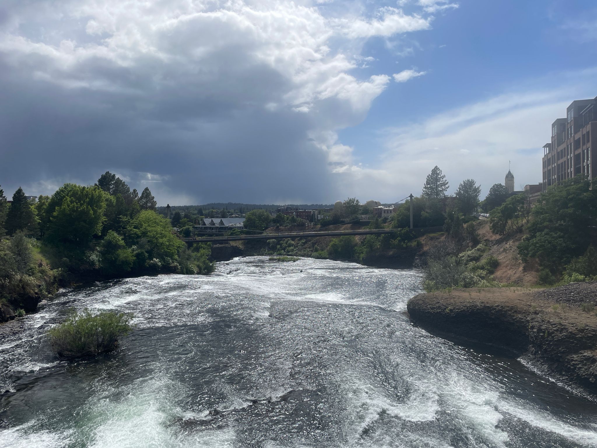 Wide river with rapids under a dramatic cloudy sky, with distant buildings. - Spokane Falls loop