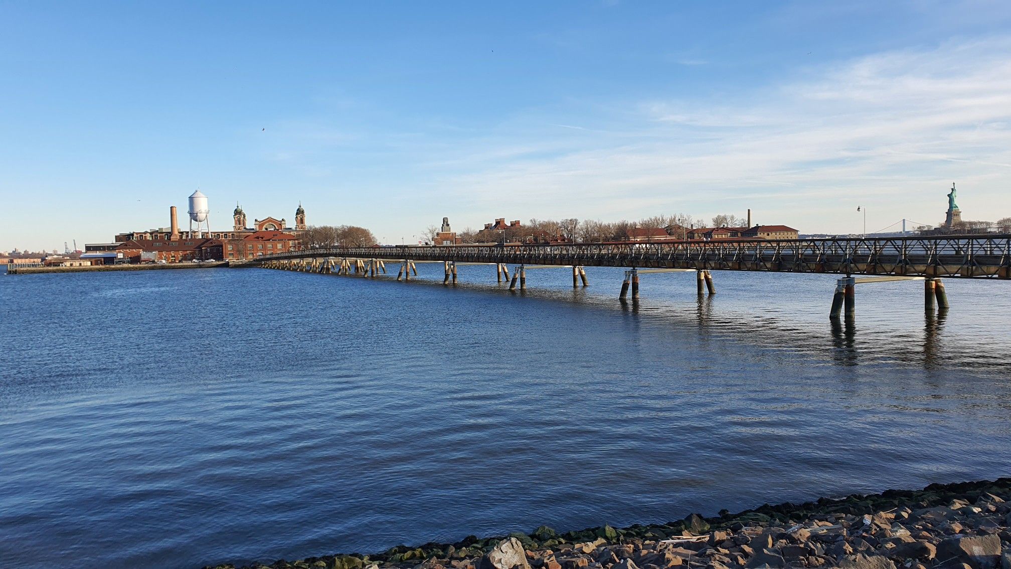 Long pier over water leading to distant buildings under a clear blue sky. - LeFrak Point Lighthouse – Liberty Landing Marina loop from 2nd Street